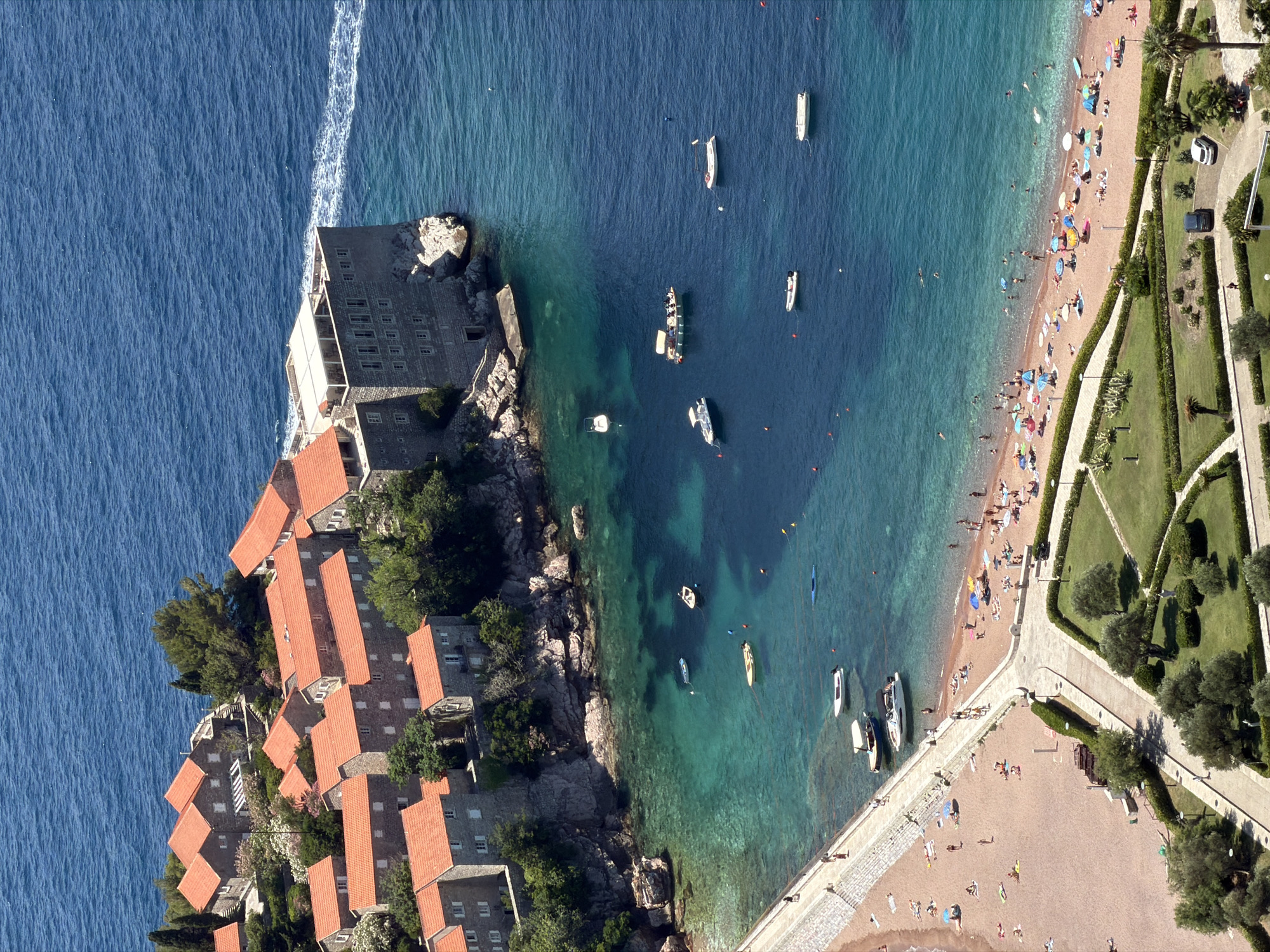 An aerial view captures the island of Sveti Stefan, Montenegro, a fortified village now a luxury resort, connected to the mainland by a narrow causeway. The island features numerous stone buildings with distinctive orange-tiled roofs, clustered tightly together on rocky terrain. A larger, imposing stone structure with numerous windows stands prominently on the easternmost point of the island.
The surrounding waters exhibit a gradient of blues and turquoises, with clear visibility into the shallow areas revealing the sandy seabed and scattered rocks. Several boats, including small motorboats and a larger vessel, are anchored or moving in the bay.
To the south of the island, a sandy beach is populated with numerous sunbathers relaxing under colorful umbrellas. A paved walkway separates the beach from the sea, and a few people are visible strolling along it. Adjacent to the beach, a well-maintained park area features manicured lawns, pathways, and scattered trees. A few parked cars are visible on a road bordering the park. The overall scene depicts a sunny day at a popular coastal resort.