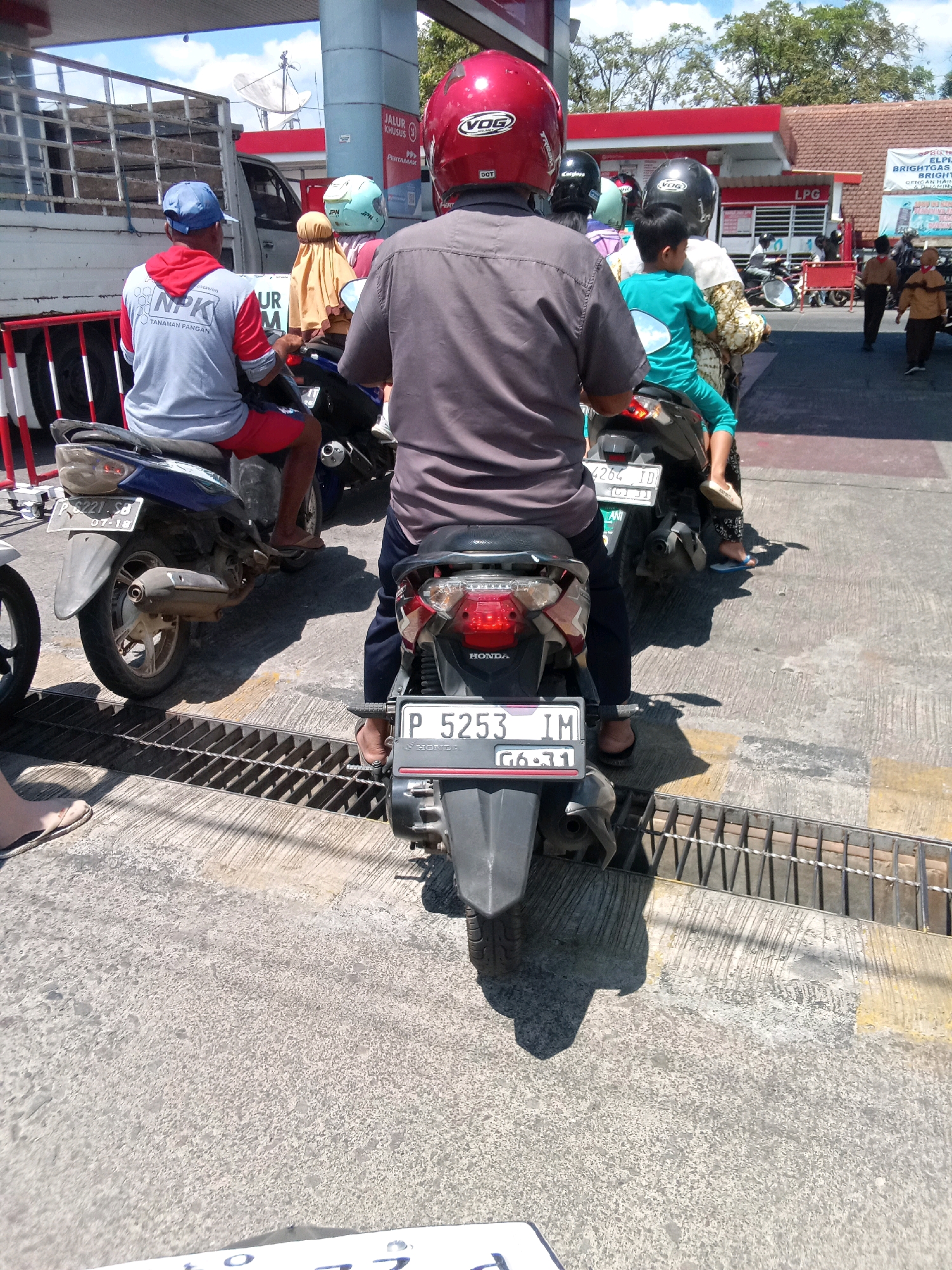 A group of people on motorcycles are queuing at a gas station in Gebang, Indonesia. The scene is outdoors, under a bright sky, with a red and white gas station building in the background. Several motorcycles are visible, some with riders wearing helmets and casual clothing. The ground is paved with concrete and has a metal grate running across it. In the foreground, a person wearing a blue cap and a gray shirt with "NPK TANAMAN PANGAN" written on it is on a blue motorcycle. Another person in a gray shirt and red helmet is on a black motorcycle behind them. A child in a green shirt is seated on the back of a motorcycle in the middle of the frame. The license plate on the motorcycle in the foreground reads "P 5253 IM". In the background, a white truck is parked, and other people and motorcycles are visible at the gas station. A satellite dish is mounted on a pole on top of the gas station building.