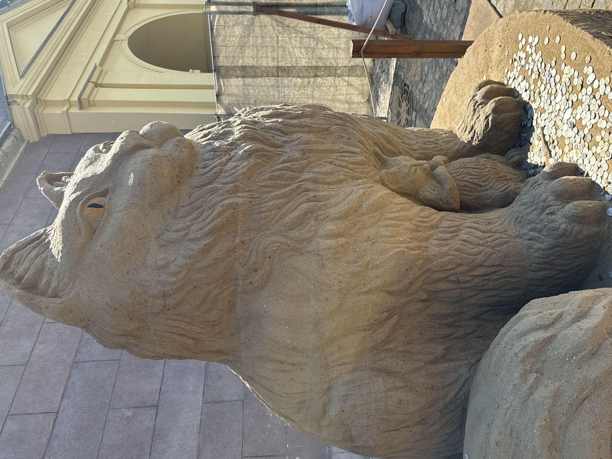 The image shows a sand sculpture of a cat with kittens in Saint Petersburg, Russia. The sculpture is detailed and decorated with coins, located in an outdoor public space with a light-colored building in the background. The sculpture is on a paved surface, and there is some construction scaffolding visible in the upper part of the frame. The focus is on the sculpture's texture and the details of its construction.