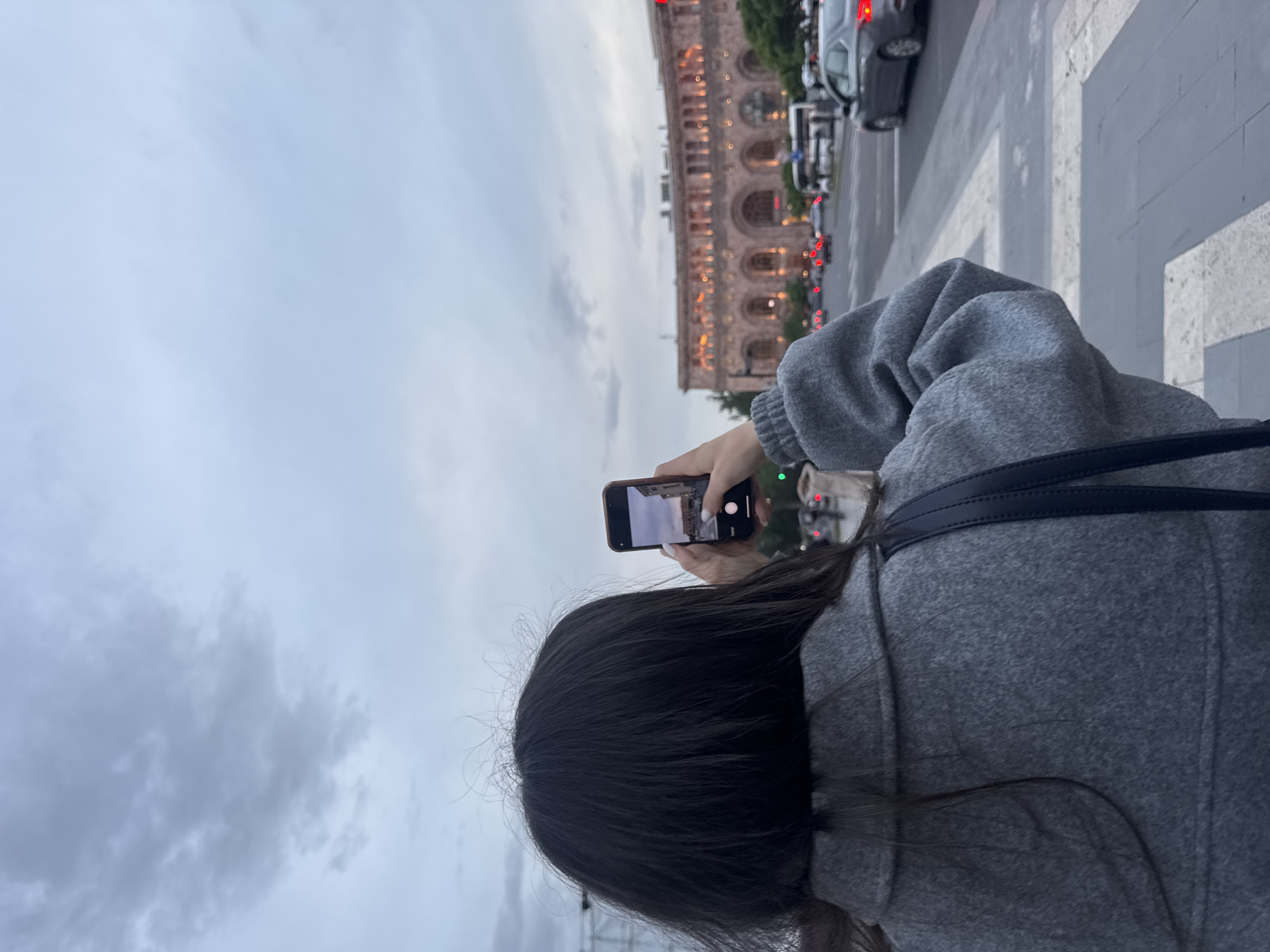 A person with dark hair, wearing a gray sweatshirt, is taking a photo with a smartphone of a building in Yerevan, Armenia. The building has multiple arched windows and appears to be lit up in the evening. Cars are visible on the street in front of the building, and the person is standing on a crosswalk. The sky is cloudy.