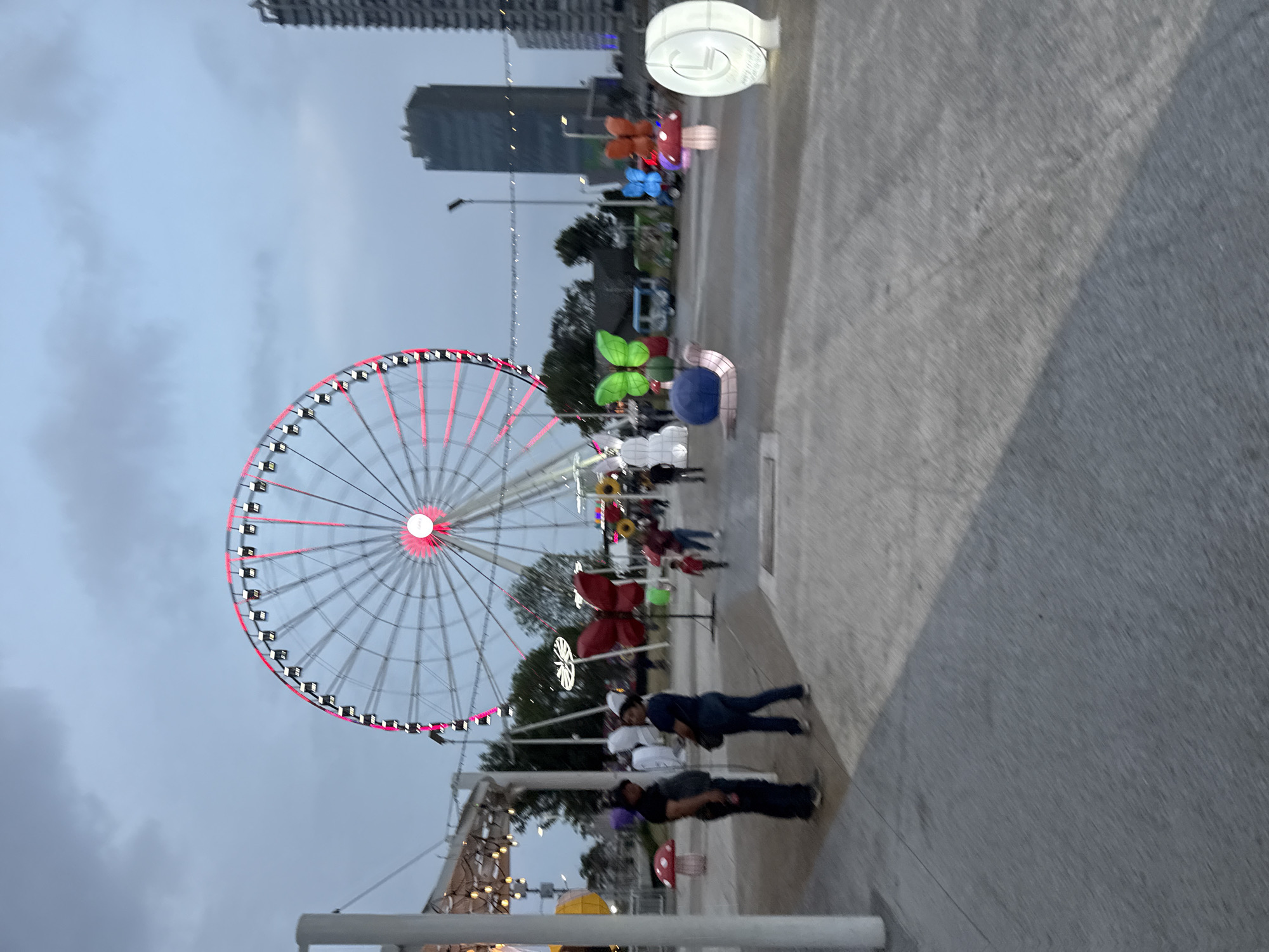 The image shows a ferris wheel in Puebla, Mexico, with a red and white illuminated frame against a grey sky. Several tall buildings are visible in the background. In the foreground, decorative lights in the shape of butterflies and mushrooms are seen, along with people walking on a grey paved area. Some people are looking at the lit-up objects.