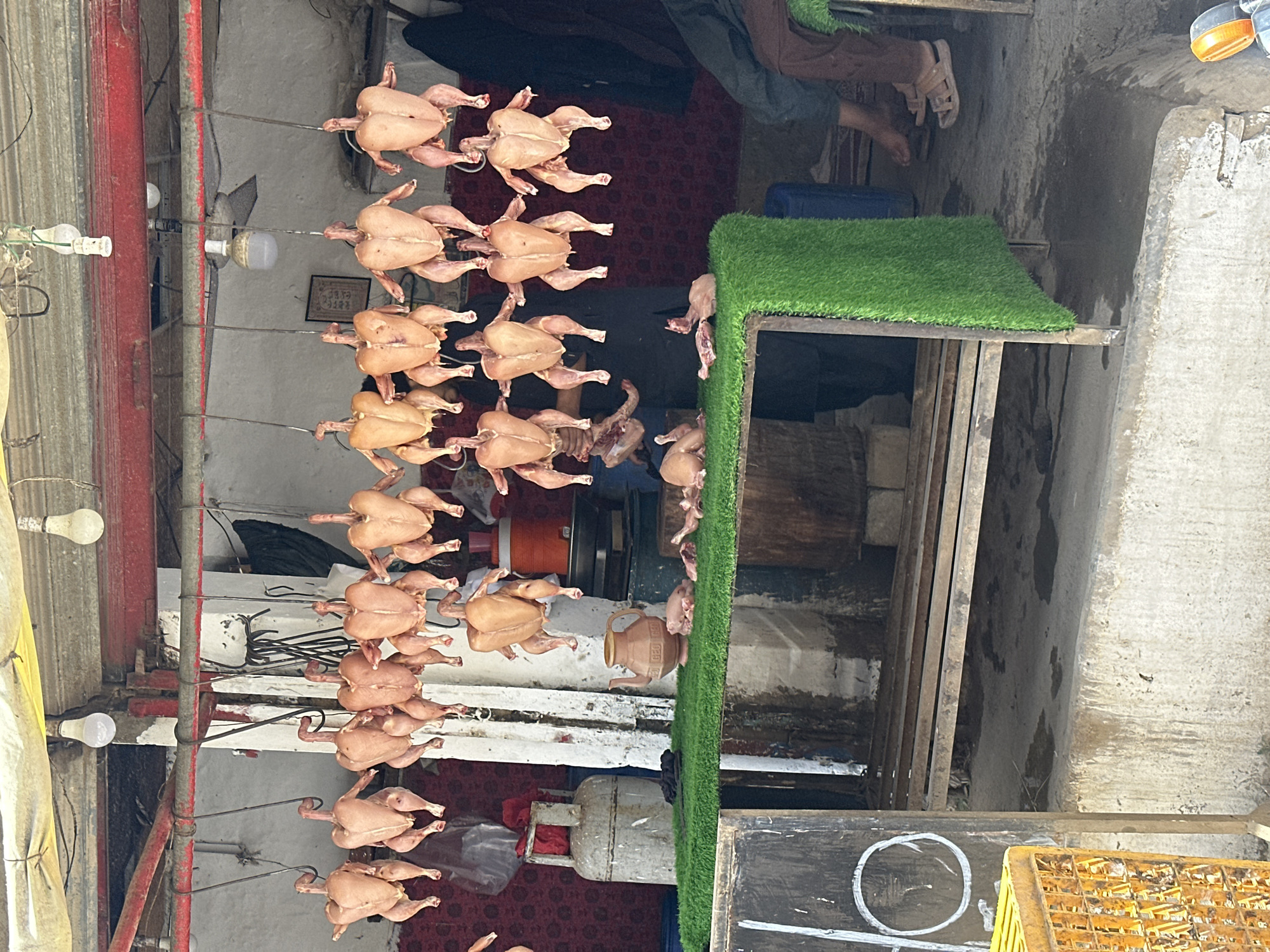 In the city of Kanozai, Pakistan, an outdoor market stall displays several whole chickens hanging by their feet from a metal bar. The chickens appear to be raw and are presented for sale. In front of the hanging chickens, a table is covered with artificial green turf, on which are displayed more chickens, some whole and some in pieces. There are also some pieces of meat on the turf. A traditional clay teapot is visible on the turf, as well as a metal gas cylinder. The stall has a corrugated metal roof supported by a wooden frame. In the background, people can be seen, some appearing to be customers and others vendors. The ground is concrete and appears to be wet in places. The overall scene depicts a bustling marketplace environment.