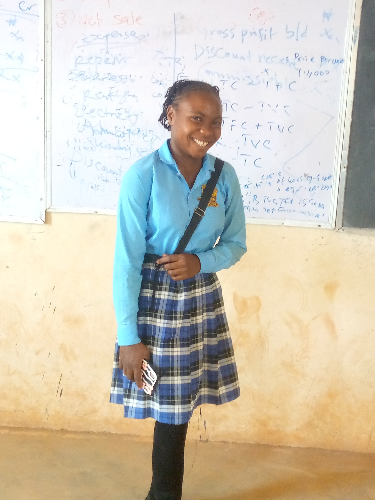 A young woman in a light blue long-sleeved shirt and a blue and black plaid skirt is smiling at the camera. She is wearing black leggings and has a black purse strap across her shoulder. She is holding a phone in her left hand. Behind her is a white board with handwritten text related to economics and accounting. The background includes a beige wall. The location is potentially a classroom in Bukuru, Nigeria, based on the context of the imagery.