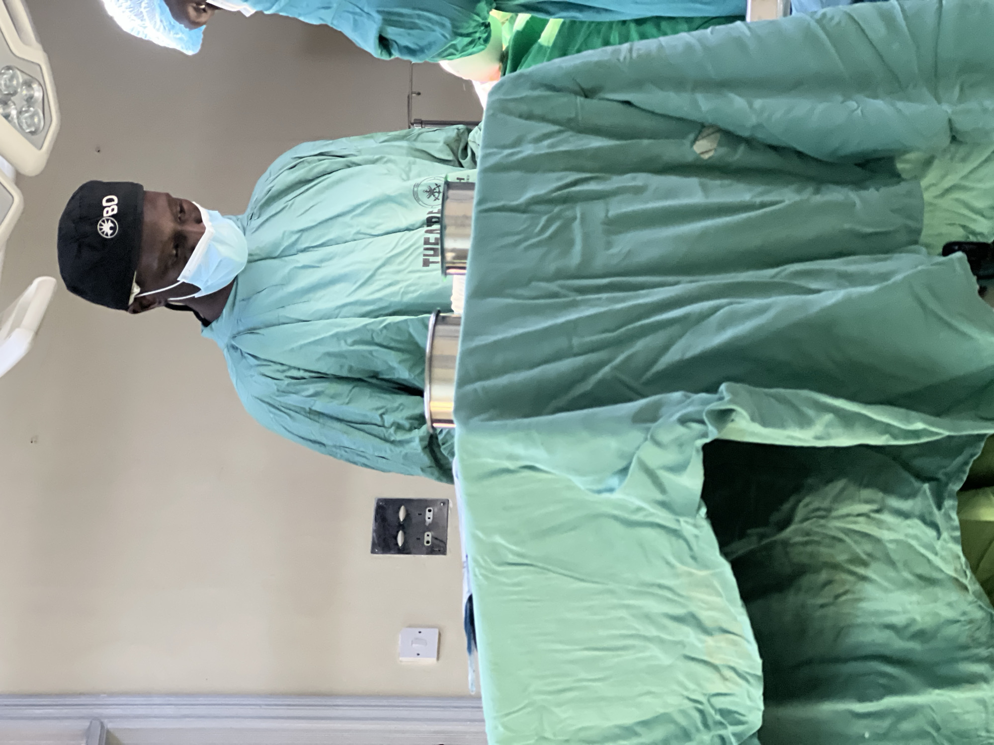 A male surgeon wearing surgical scrubs, a surgical cap with "BD" written on it, and a surgical mask is shown. He is standing in an operating room, in what could be a hospital setting, possibly in Nairobi, Kenya. He is positioned beside a surgical table draped with green fabric. An overhead surgical light is visible. The wall behind him is a light beige color. Electrical outlets are on the wall, along with what appears to be a light switch.