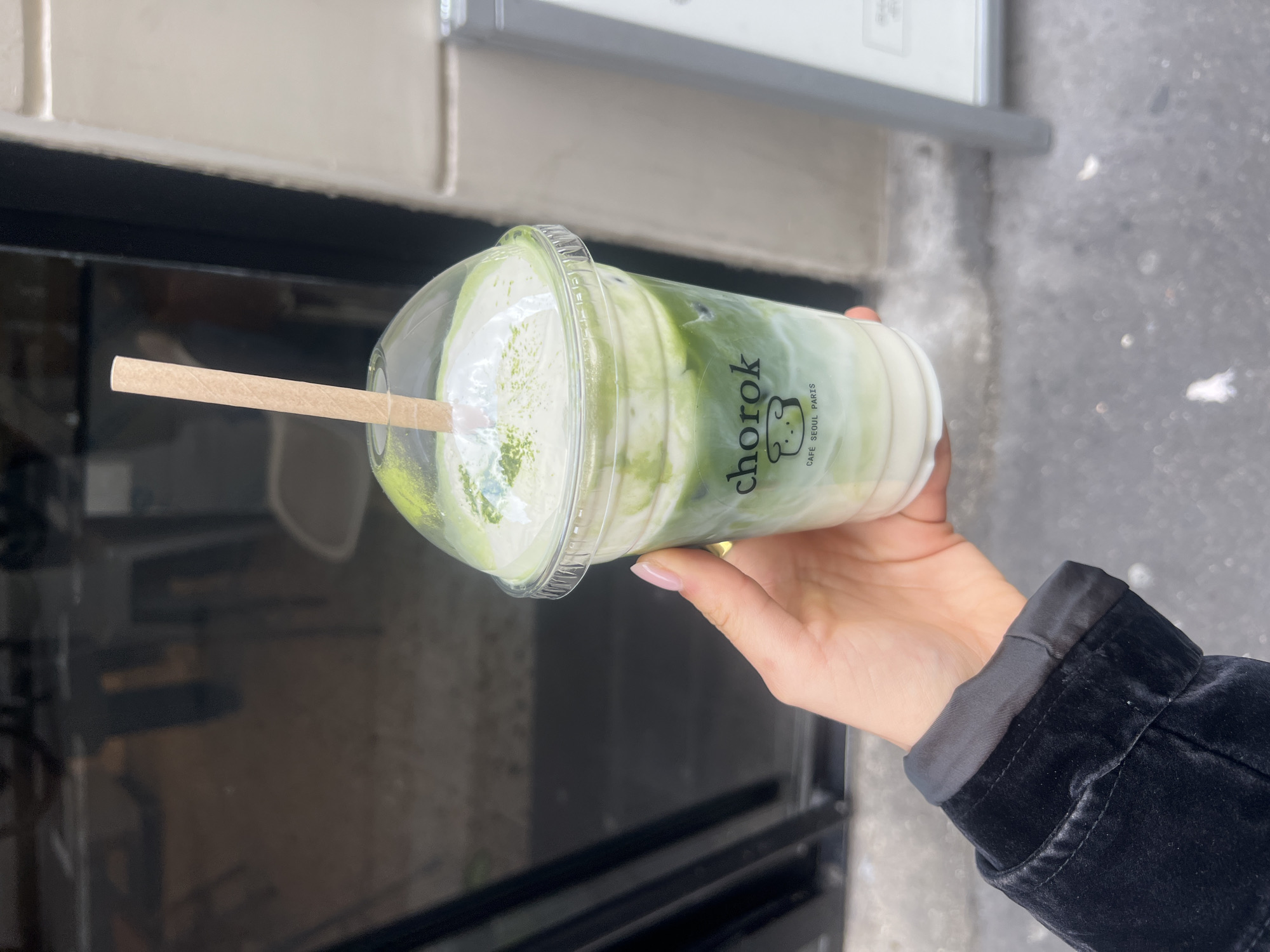 A person holds a plastic cup containing a layered green and white drink with a straw in their hand. The cup is labeled "chorok" with the words "CAFE SEOUL PARIS" beneath it. The person wears a black jacket. The background appears to be an urban setting, possibly in Paris, France. A dark surface, possibly a storefront window or counter, is visible behind the cup.