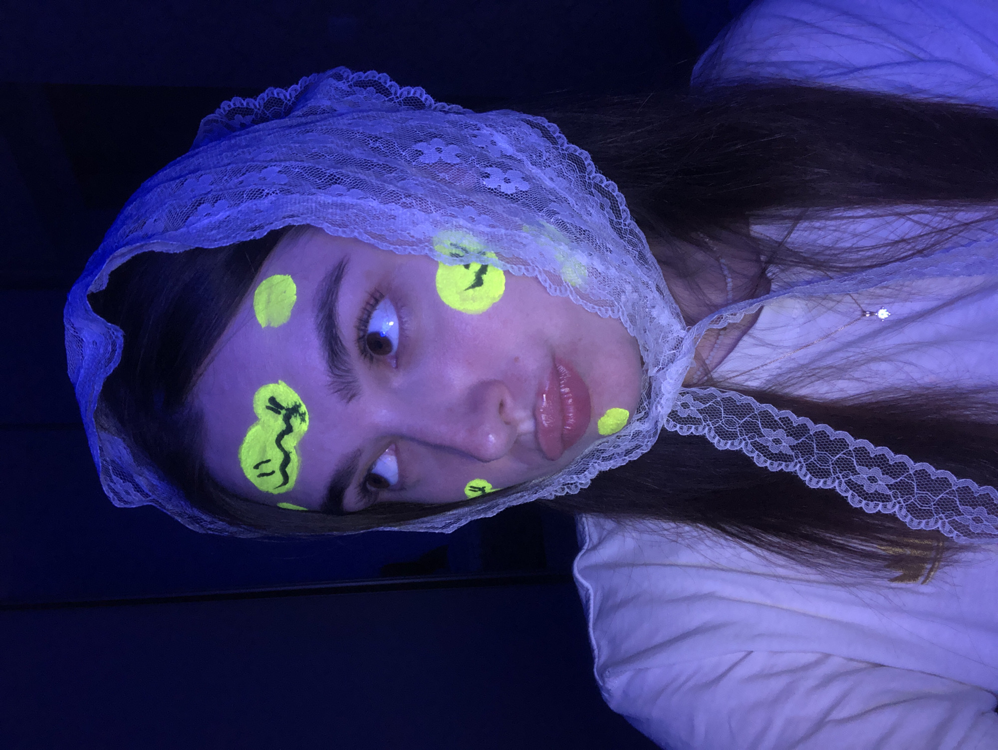 The image shows a young woman in a dimly lit room, possibly an indoor setting within the city of Noviy, Russia. She is wearing a white t-shirt and a white lace headscarf. Her face is adorned with fluorescent yellow markings, including two circular shapes on her cheeks, one on her forehead, and one near her eyebrow. The markings on her forehead and right cheek appear to be stylized smiley faces. The lighting in the room is predominantly blue, suggesting the presence of a blacklight, which causes the yellow markings to glow. Her expression is contemplative as she looks away from the camera, with her lips slightly pursed. The background is dark and out of focus.