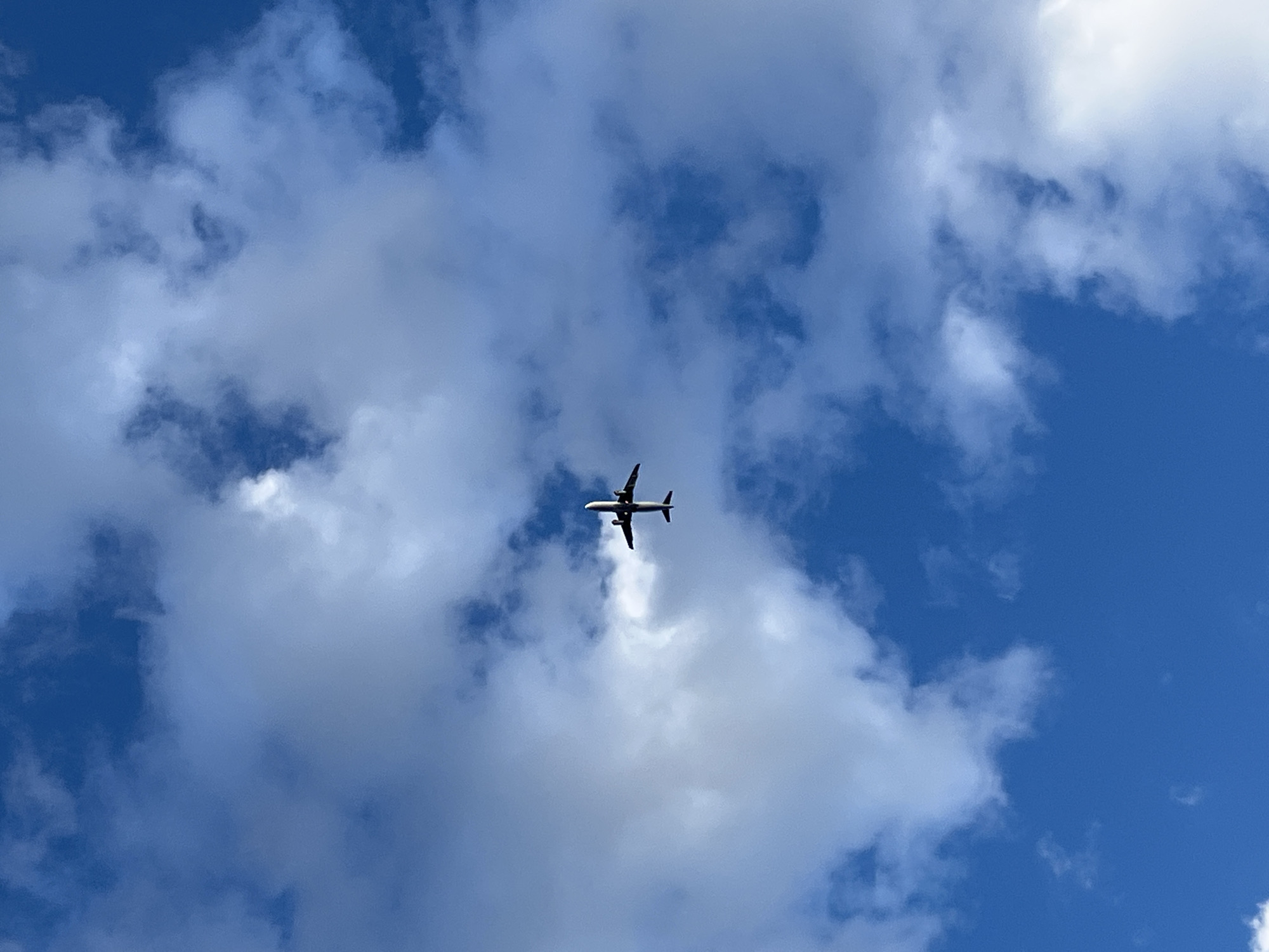 A silhouetted airplane is captured from below against a bright blue sky filled with scattered white clouds. The aircraft is in mid-flight, with its wings extended and its fuselage and tail clearly visible against the lighter background of the clouds. The perspective is looking directly upwards, emphasizing the vastness of the sky and the distant position of the plane. The scene depicts a common aerial view, with no people or structures other than the aircraft itself visible.