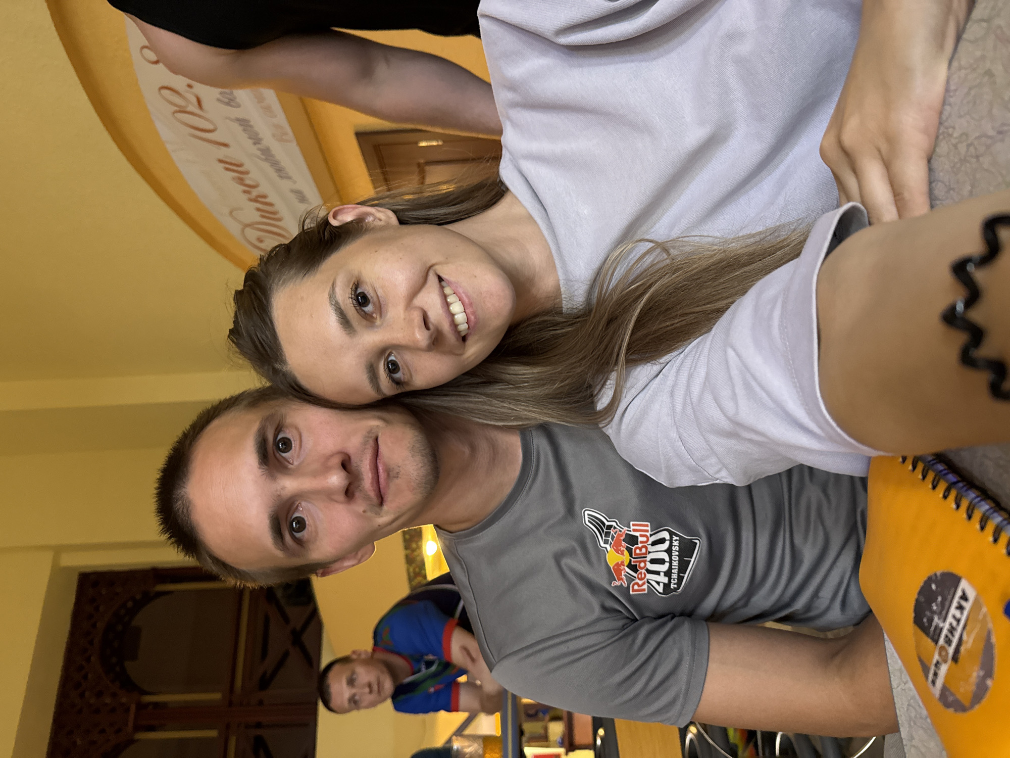 In a selfie taken from a low angle, a man and a woman are seated at a table. The man, on the left, has short brown hair, a prominent forehead, and light brown eyes. He wears a grey t-shirt with a Red Bull 400 Tchaikovsky logo. The woman, on the right, has long blonde hair and a bright smile, wearing a light purple sweatshirt. Behind them, another man in a blue and red shirt is visible, as is a partial view of a black shirt belonging to a third person. A yellow notebook with a circular logo is on the table in the foreground. The background features a yellow wall with ornate wooden architectural elements and a sign with Cyrillic lettering that reads "Дворец 102." The setting appears to be indoors, possibly a cafe or community center, in the city of Chaikovsky, Russia, suggested by the event logo. The overall atmosphere is casual and personal.