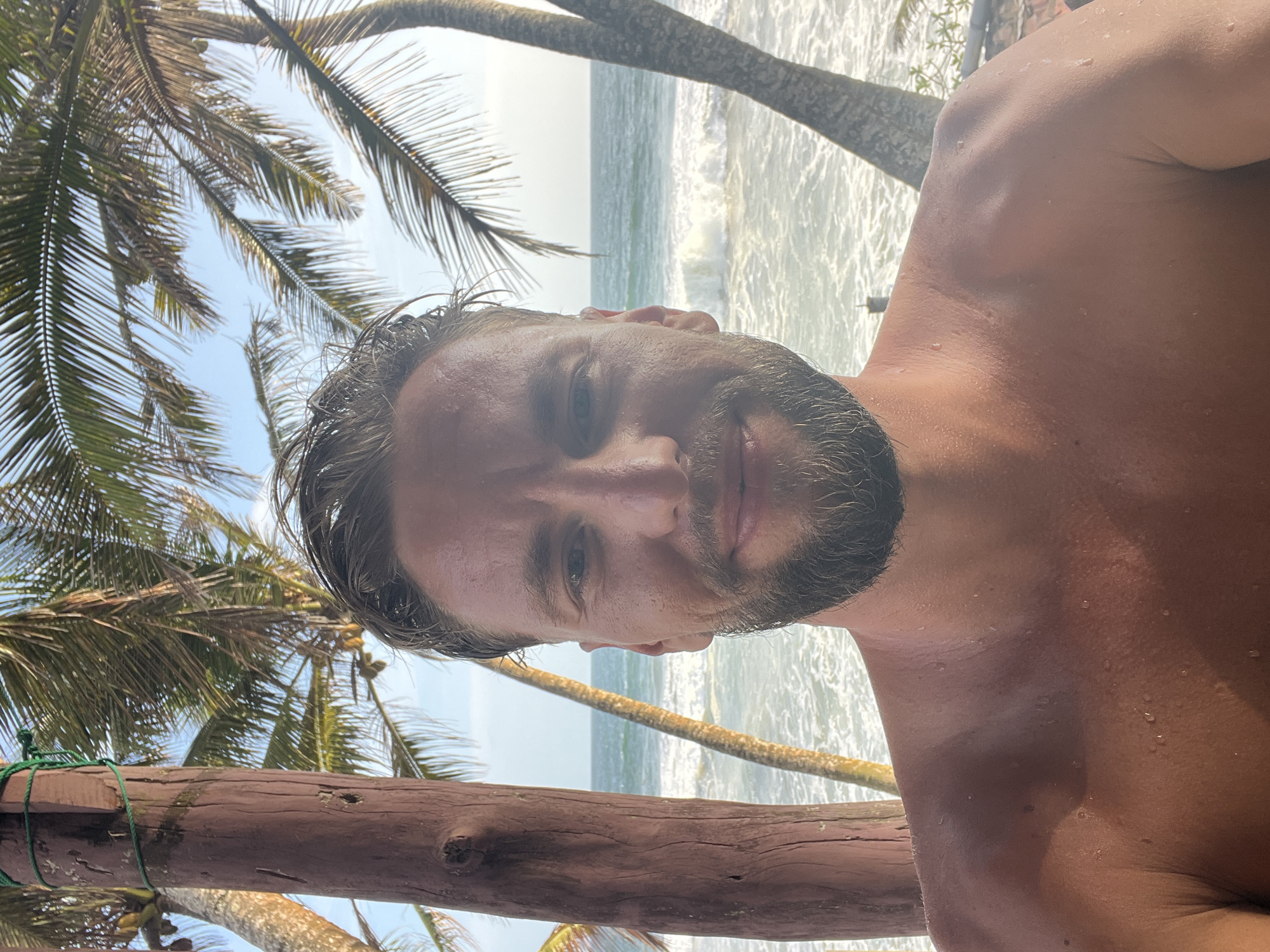 A selfie taken from the chest up shows a shirtless man with wet hair and a beard. He is standing outdoors, with palm trees and the ocean visible behind him. The man is smiling slightly. The environment appears to be a beach in Rajgama, Sri Lanka, with the ocean providing a backdrop. The lighting suggests it is daytime, possibly late afternoon. There is a wooden post to the left of the man.