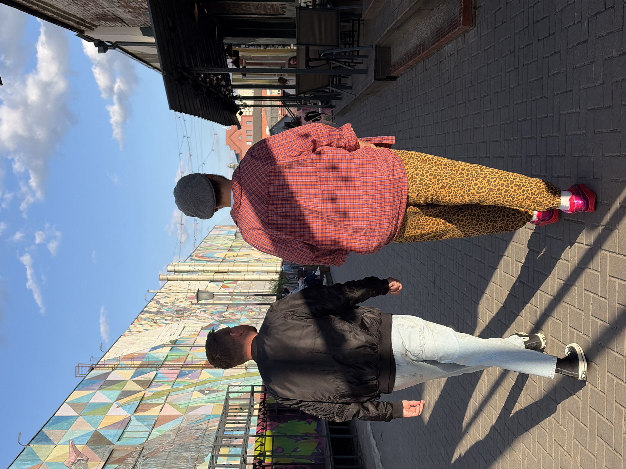 Two people are walking outdoors along a brick-paved pathway. The person closer to the camera is wearing a black bomber jacket, light blue jeans, and black and white sneakers. The second person is wearing a gray newsboy cap, a red plaid shirt, leopard print pants, and pink and red platform shoes. They are walking past a building with a large, colorful geometric mural. The location is in Minsk, Belarus. Above them, there is a blue sky with scattered white clouds. A building with awnings and outdoor seating is visible in the background. Harsh shadows are cast across the brick pavement, indicating bright sunlight.