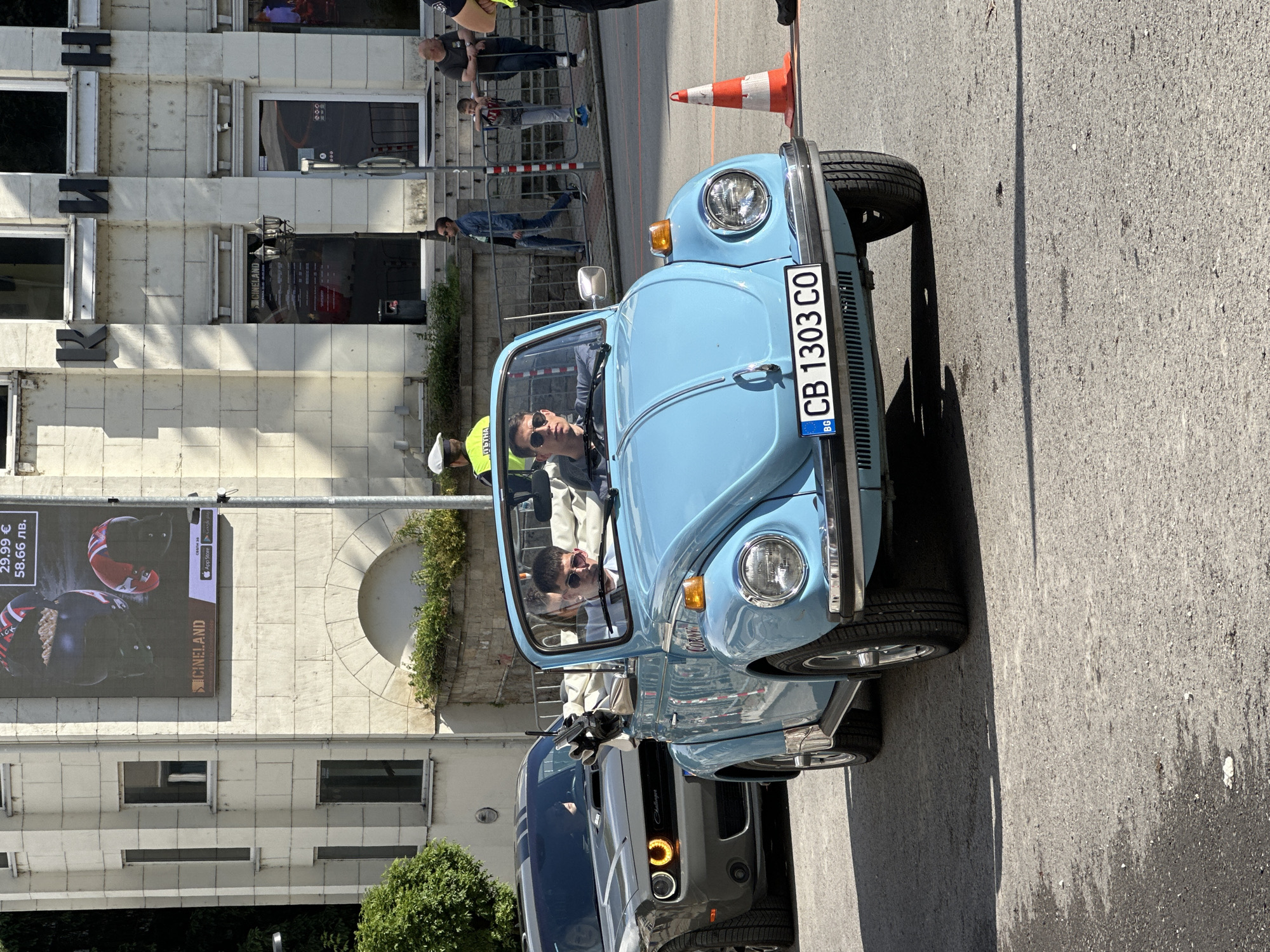 The image captures a scene in Veliko Tarnovo, Bulgaria, showcasing a blue Volkswagen Beetle convertible parked on a city street. Inside the car, two individuals are visible. The driver is wearing sunglasses and a light-colored jacket. The car's license plate reads "CB 1303 CO." Behind the Beetle, a gray Dodge Challenger is partially visible.
The background features a building with an advertisement banner for "CINELAND" displaying the price of "29.99 €" and "58.66 лв.". The building's facade also exhibits signage with large, dark-colored letters "K," "H," and "И." On the street, there is an orange traffic cone near the Beetle. Several pedestrians can be seen observing the scene.
