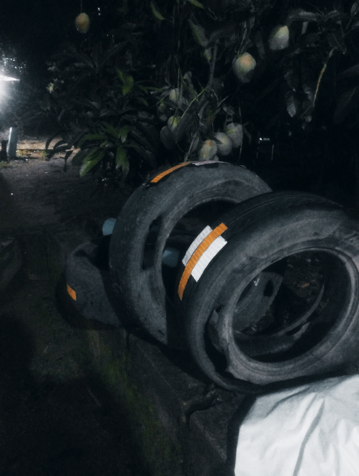 The image depicts an outdoor scene at night, likely in Jos, Nigeria, with two tires prominently featured in the foreground. The tires are dark and appear weathered, with orange and white reflective tape affixed to their sides. One tire is positioned slightly behind and above the other, creating a layered effect. In the background, a tree with visible fruits, possibly mangoes, is present. The lighting suggests a low-light environment, with a bright light source visible on the left side of the frame, illuminating a portion of the ground and foliage. The ground appears to be dirt or gravel, with some green foliage and darker shadows creating depth. A white cloth or sheet is draped in the lower right corner, partially obscuring the ground. The overall atmosphere is dim and somewhat rustic.