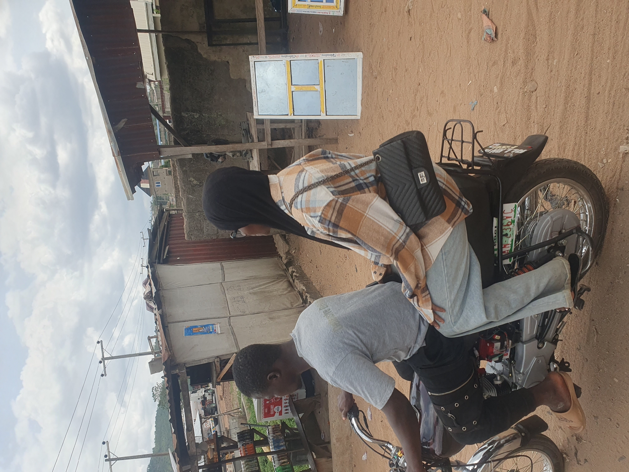 Two people are on a motorcycle. The driver, a man, is wearing a gray shirt and black pants with silver accents. The passenger, a woman, is wearing a black hijab, a plaid shirt, and light-colored pants. The motorcycle is black with chrome accents.
The background suggests an urban environment, potentially a street in Lokoja, Nigeria. There are buildings, possibly shops or homes, visible. A small business selling beverages and snacks is visible. The ground appears to be dirt or gravel.