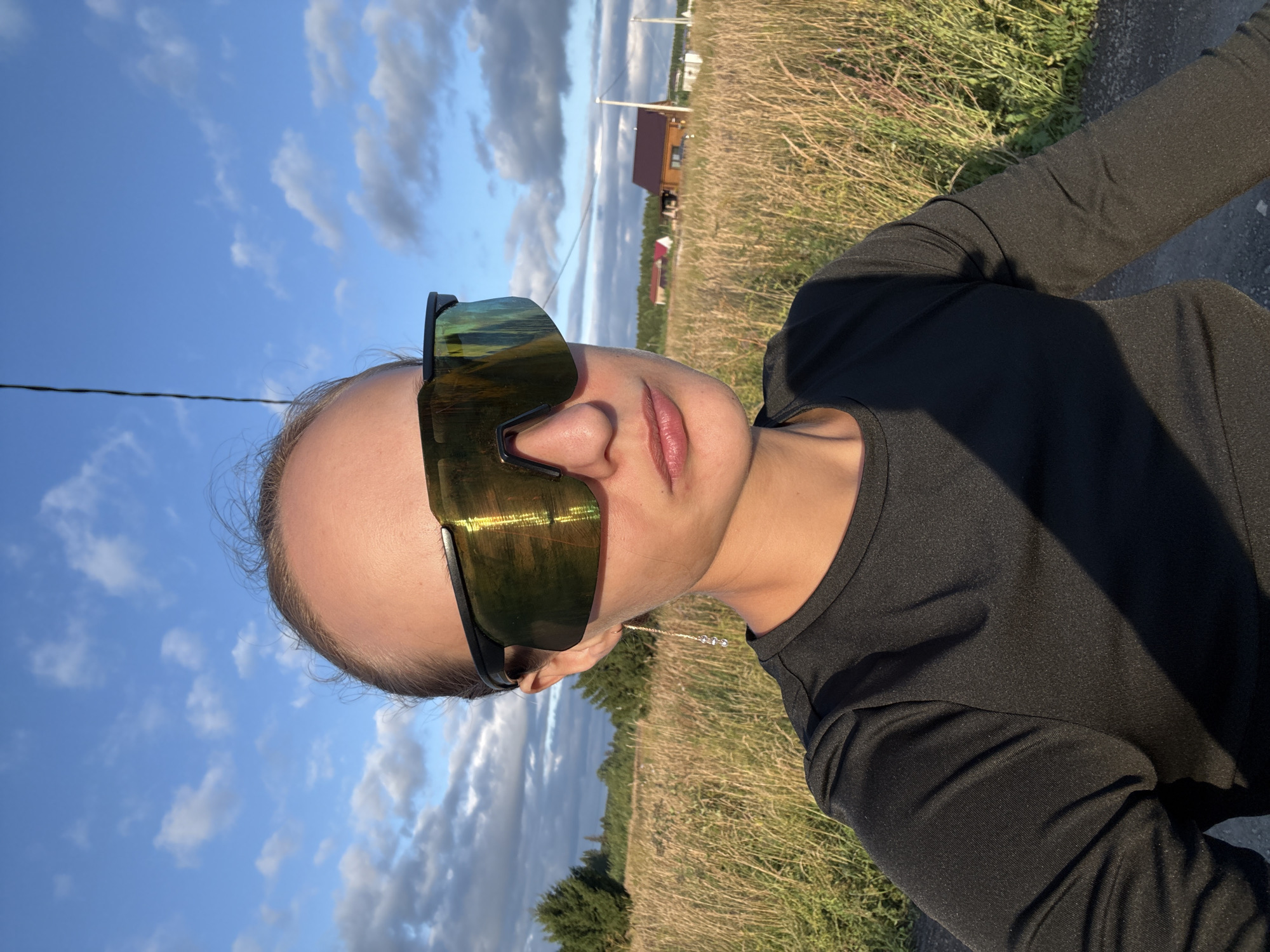 A medium shot captures a woman from the chest up, wearing large, reflective wrap-around sunglasses. She has fair skin and her hair is pulled back, with some strands loose around her face. She wears a black long-sleeved shirt. A single gold earring with three small clear beads hangs from her left ear. The background shows a field of dry grass under a bright blue sky with scattered clouds. In the distance, several houses with red roofs are visible, along with trees and utility poles. The image appears to have been taken outdoors in a rural area, possibly in the vicinity of Podshivalovo, Russia, given the architecture and landscape.