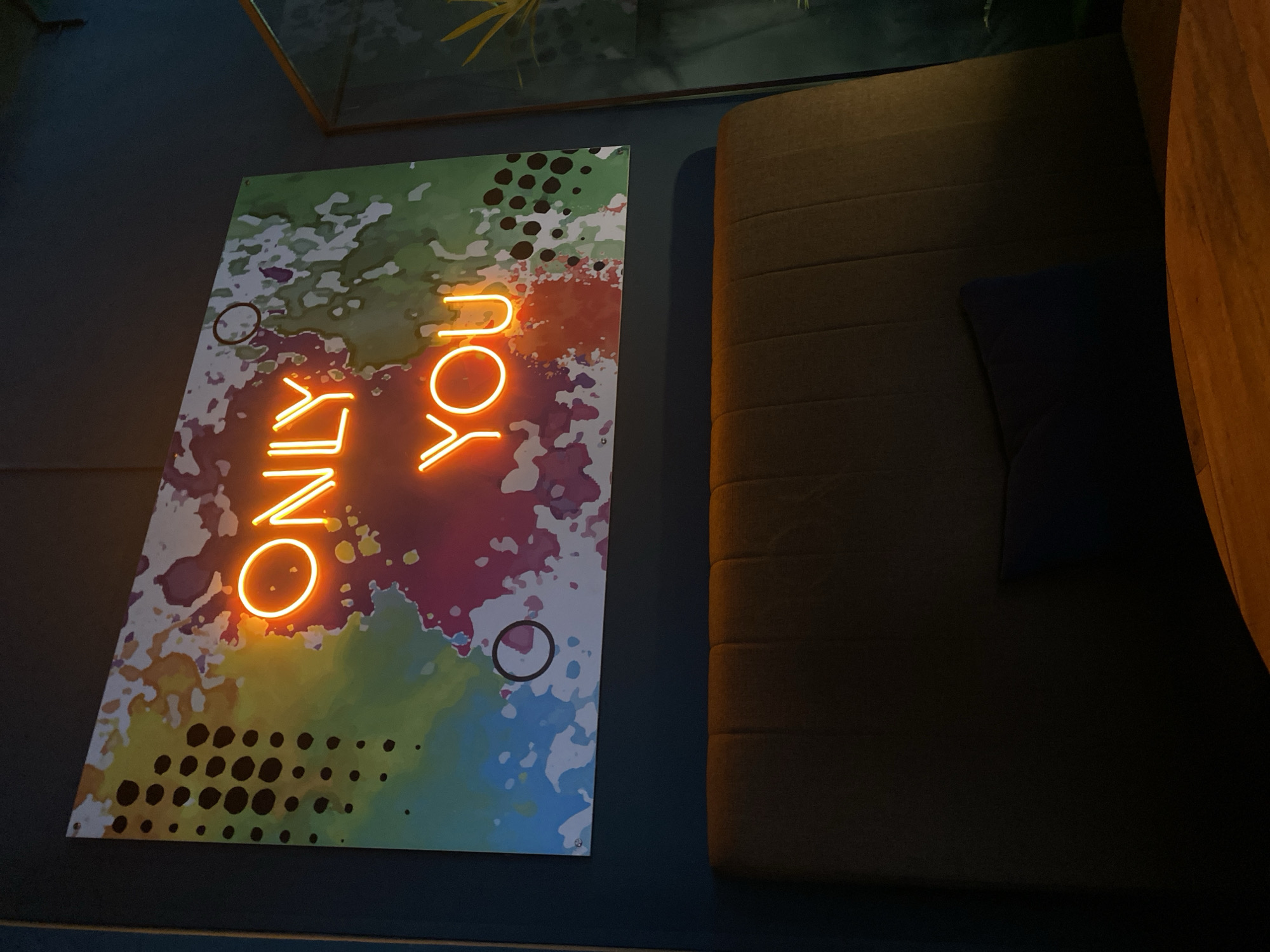 A close-up shot displays a wall with decor. A vertically oriented, rectangular print features a multicolored, watercolor-esque design with the words "ONLY YOU" in neon orange lettering. The print is positioned left of center. Adjacent to the print, to the right, is a tufted, brownish-gray couch with a dark blue cushion. The couch extends beyond the frame on the right. Above the print, a glass-encased display is partially visible. The wall is a dark, muted blue color. The scene is dimly lit. There is no indication of the location being Chaikovsky, Russia.