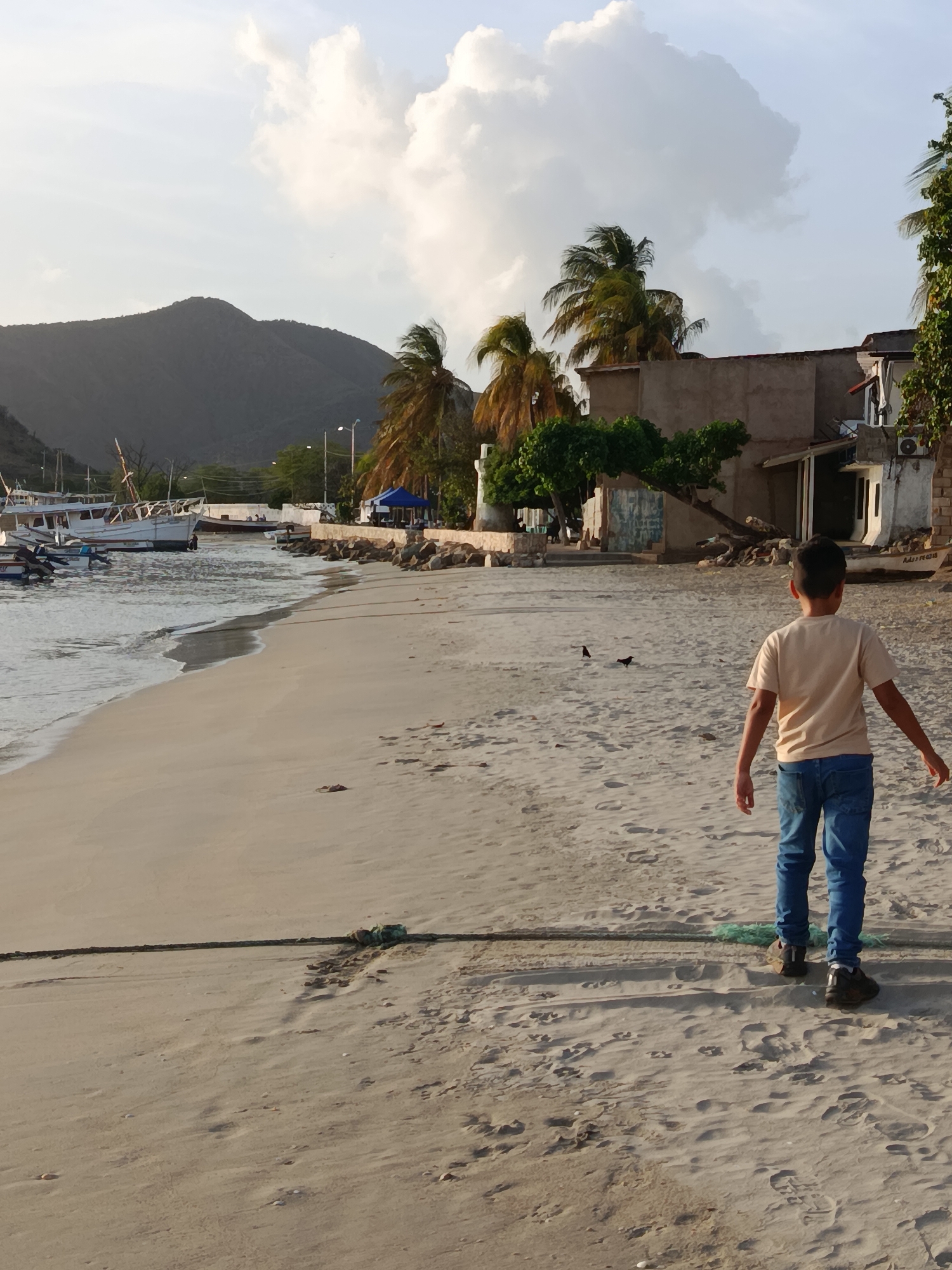 A young boy walks on a sandy beach towards the right of the frame in Juan Griego, Venezuela. The boy, viewed from behind, wears a light-colored t-shirt and blue jeans. He is barefoot. The beach stretches into the distance, meeting the calm ocean on the left. Several boats are moored in the water near the shore, and a rocky embankment separates the beach from a built-up area with buildings and palm trees in the background. A mountain range is visible in the far distance. The sky is partly cloudy, with a large white cloud dominating the upper right portion of the frame. Two birds are visible on the sand in the middle ground. The lighting suggests it is late afternoon or early evening.