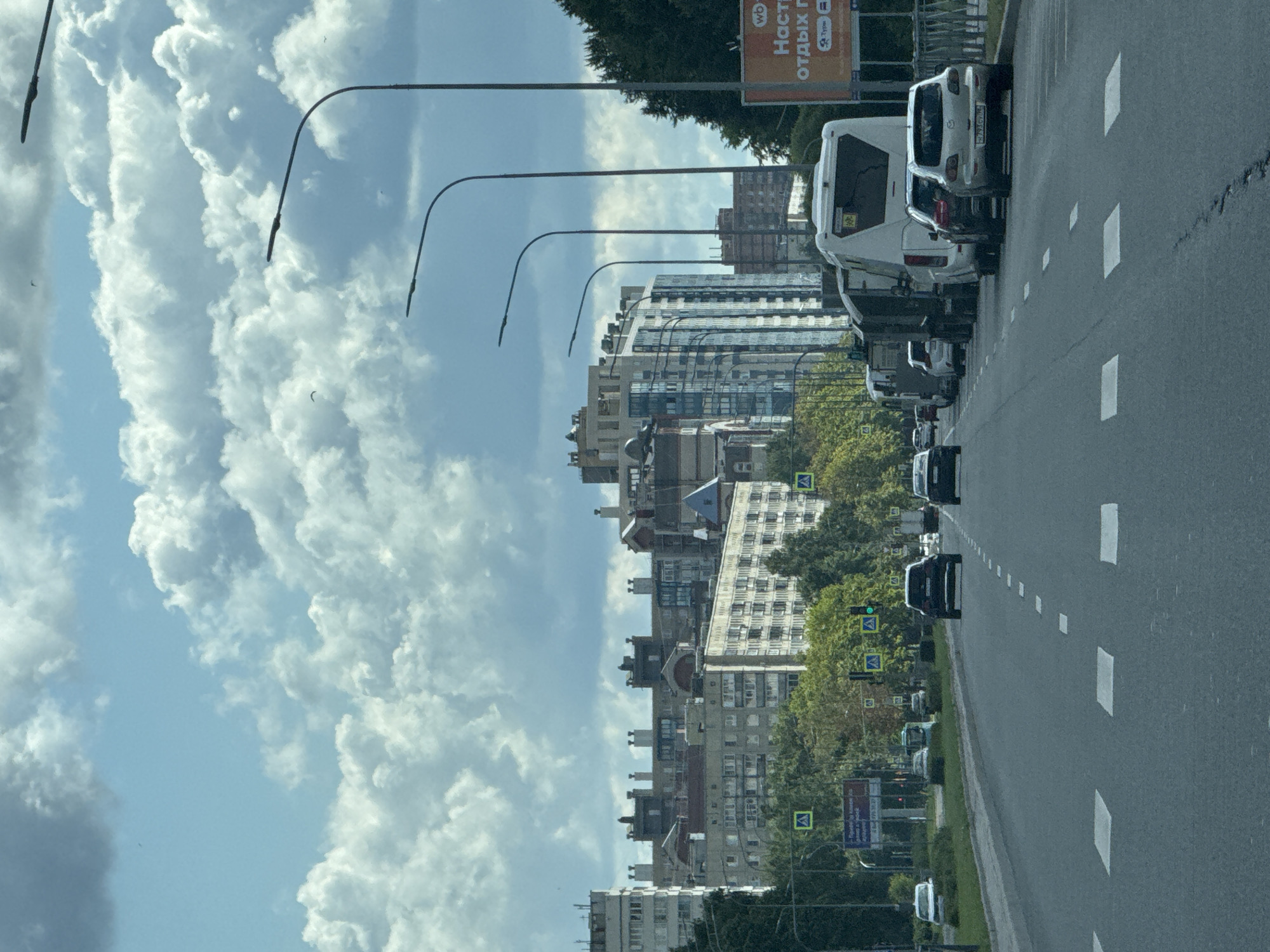 The image depicts a street scene in Sinyavino, Russia, under a partly cloudy sky. A multi-lane asphalt road occupies the foreground, with white dashed lines and white square pedestrian crossings marking the lanes. Several vehicles are present: a silver Mazda sedan with a license plate ending in "0905" is parked on the right shoulder, a dark SUV, a white bus, and other vehicles are in traffic lanes. A billboard advertising "Настя отдых п" is visible on the right side of the road, partially obscured by trees. In the background, a row of multi-story residential buildings and a modern glass-fronted building are situated behind a line of trees. Streetlights with curved arms line the right side of the road. The overall environment suggests an urban or suburban setting within Sinyavino.