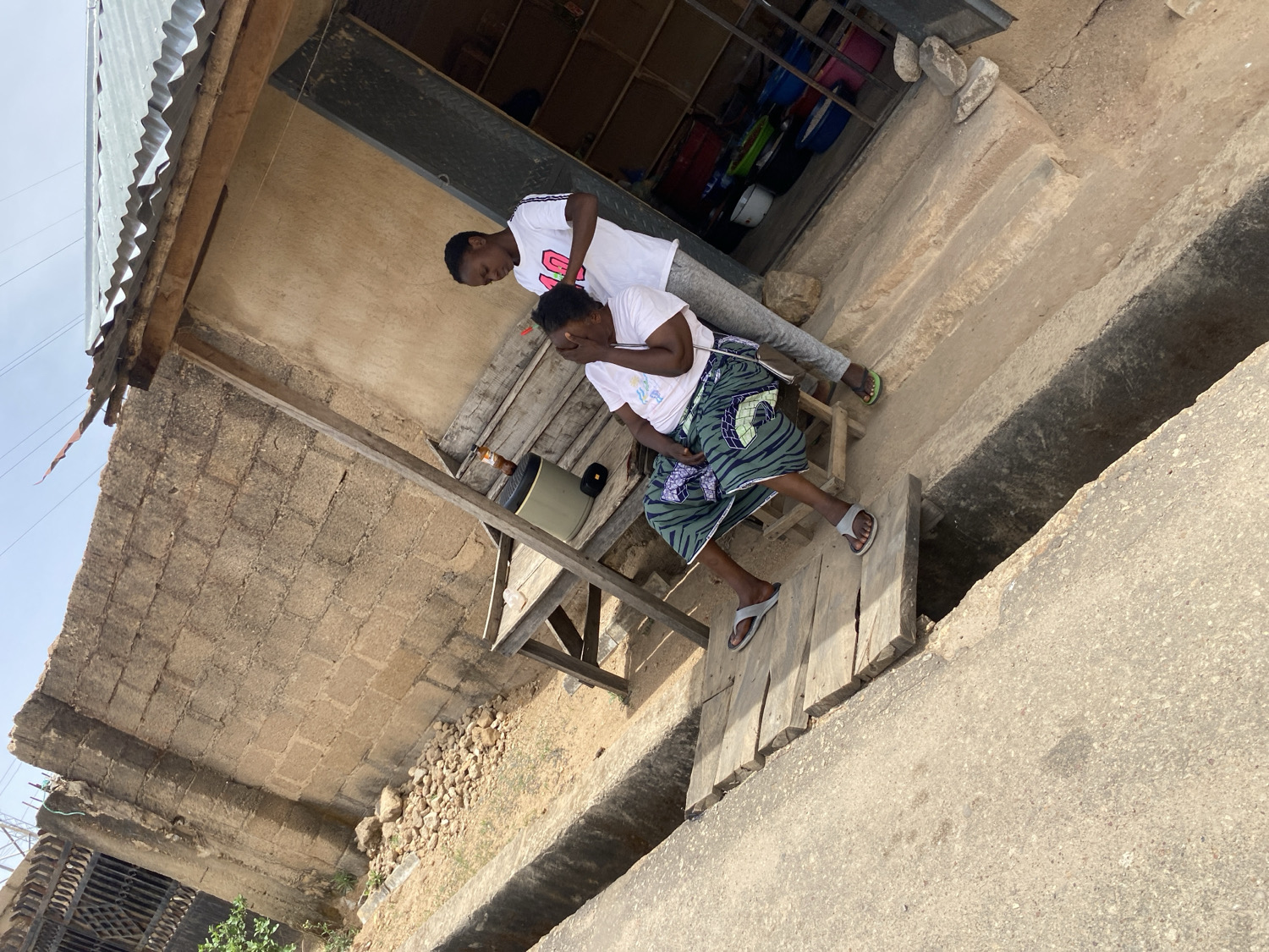 This eye-level outdoor photograph captures a candid moment between two individuals in what appears to be a street or informal market setting in Jos, Nigeria, during bright daylight.
In the midground, a young person stands on the left, wearing a white t-shirt with the word "LOVE" (or similar) visible on the chest, grey trousers, and green flip-flops. They are looking down at another person who is seated. This second individual, likely female, is positioned on a low, rustic wooden stool or bench on the right. She wears a white t-shirt, a distinctively patterned green and black wrapper skirt, and grey flip-flops. Her hands are covering her face, conveying a sense of distress, sorrow, or deep contemplation. Adjacent to her on the wooden structure, a portable speaker or radio and a brown bottle are visible.
The scene is set against a simple building constructed from cinder blocks, topped with a corrugated metal roof that extends significantly. Through an opening in the building behind the standing person, various colorful plastic containers can be glimpsed, suggesting a small shop or storage area. The ground in the foreground consists of concrete, featuring a deep, open concrete drainage ditch or gutter running diagonally. A few wooden planks form a makeshift bridge or pathway over this ditch. The sky above is bright and clear, indicating a sunny day, likely midday or afternoon. The overall impression is one of emotional vulnerability captured within an everyday, bustling environment characteristic of the specified location.
