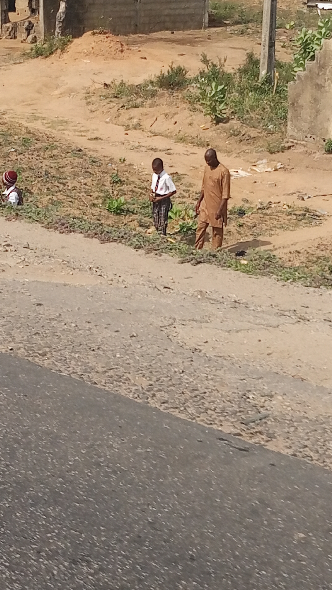 In Lokoja, Nigeria, the image shows three people next to a road. One individual is an adult male wearing brown attire. Another is a young boy dressed in a white shirt and checkered pants, suggesting a school uniform. A third, smaller individual is also present. The scene features a partially paved road alongside a dirt area with sparse vegetation and structures, suggesting a peri-urban environment.