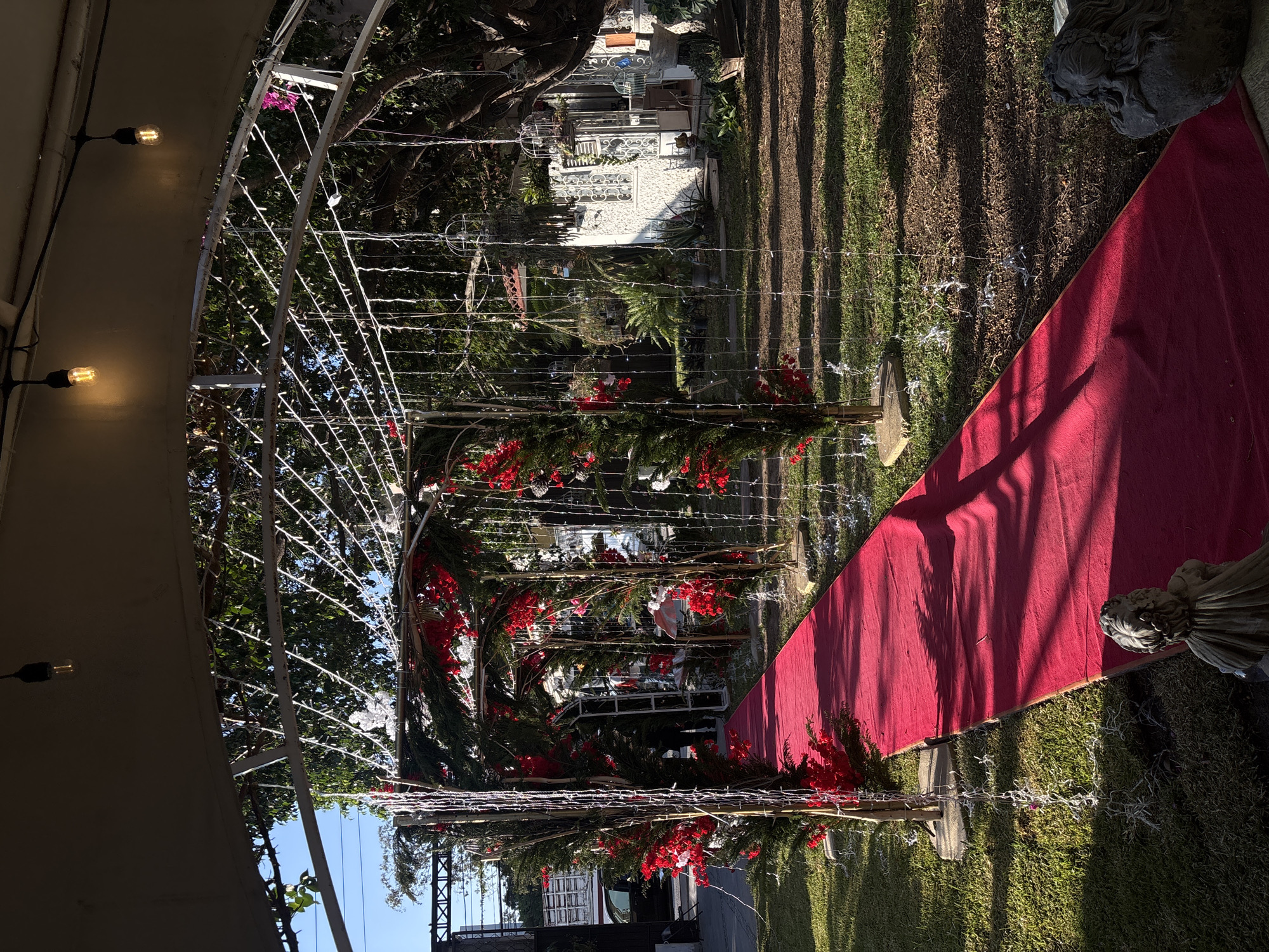 The image depicts an outdoor event space in Chilpancingo, Mexico. A red carpet runs through the center of a grassy area, flanked by decorative elements, including a metal archway adorned with red flowers and greenery. String lights are hung around the archway and along the ground. A stone sculpture sits beside the red carpet. Beyond the archway, a white building is visible in the background, along with trees and other greenery. The scene suggests a wedding or special event setup.