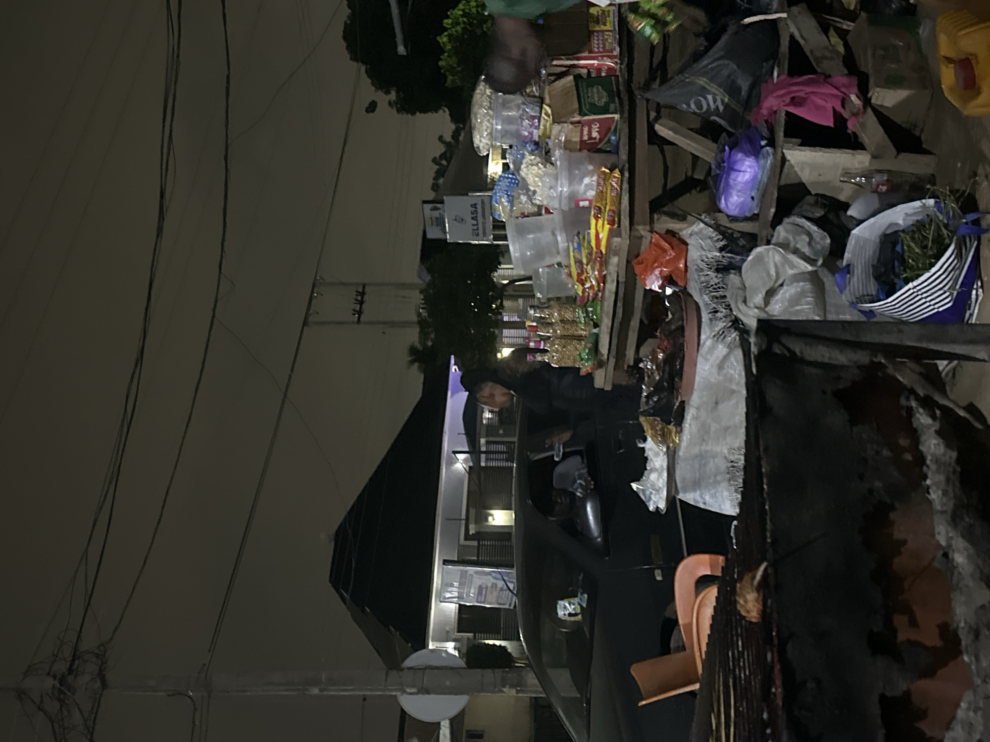 This is a nighttime, low-light, street-level photograph taken in Abuja, Nigeria. The scene depicts a roadside stall or market stand laden with various packaged goods, snacks, drinks, and what appears to be food items. The stall is constructed from wood and appears somewhat makeshift. In the background, a dark-colored car is parked, with its driver's side window partially down. A person is leaning into the car window from the driver's side, seemingly interacting with the occupant. To the left of the car, an orange plastic chair is visible. Further in the background, there is a modern-looking building with a dark pitched roof and illuminated windows, suggesting it is a residence or commercial establishment. Electrical wires and poles are prominent in the sky above, indicative of a typical urban setting. The overall atmosphere is dimly lit, with artificial lights illuminating the stall and surrounding areas.