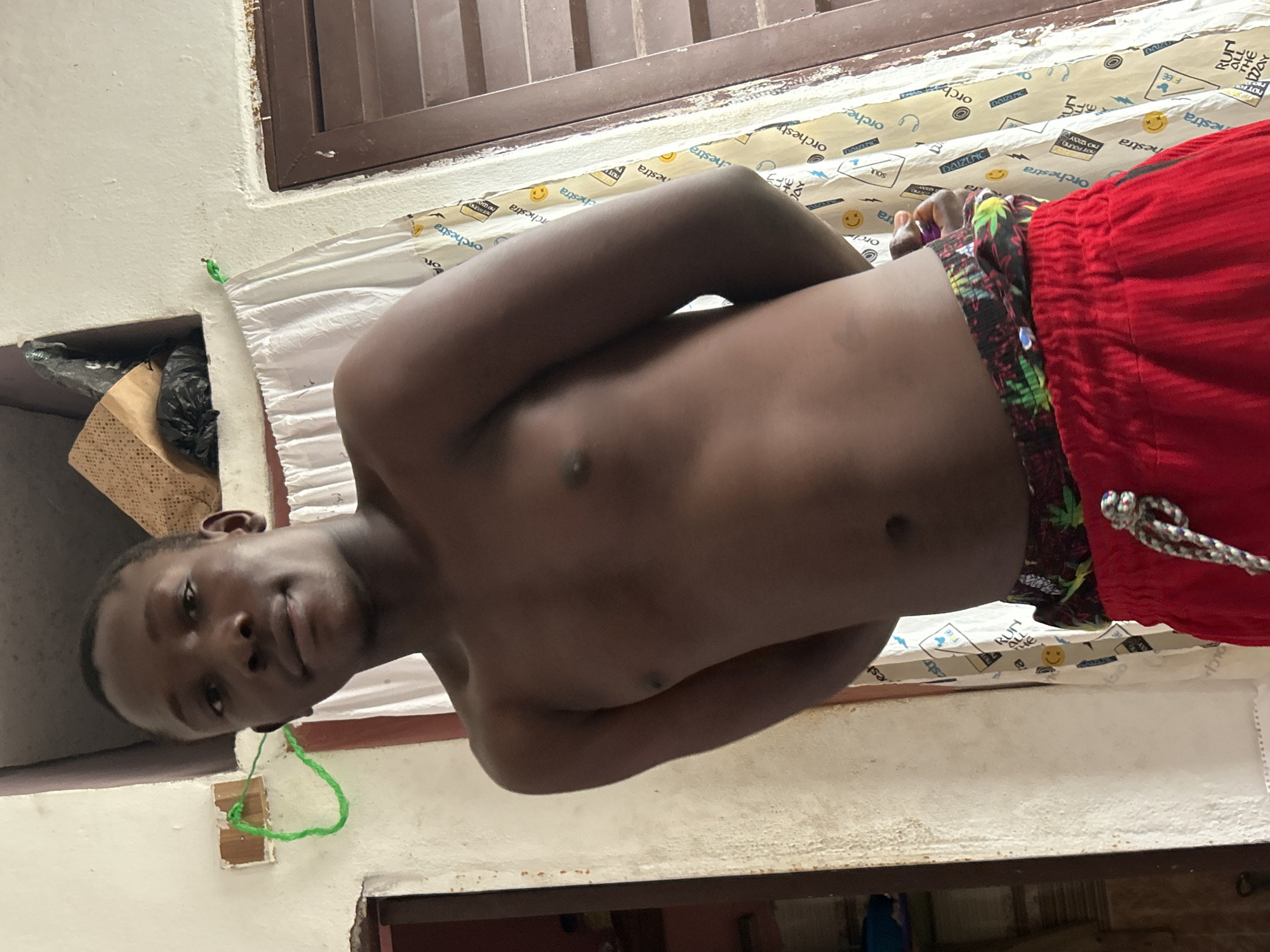 A young Black male is standing shirtless and facing the camera, his arms behind his back. He is wearing red shorts with a colorful patterned waistband. The background appears to be an indoor setting with white walls and a window covered by a white curtain adorned with repeating text and smiley face graphics. To the left, a dark doorway is visible. On the wall, a bright green string is attached to a white fixture. The image is taken in Ihiagwa, Nigeria.