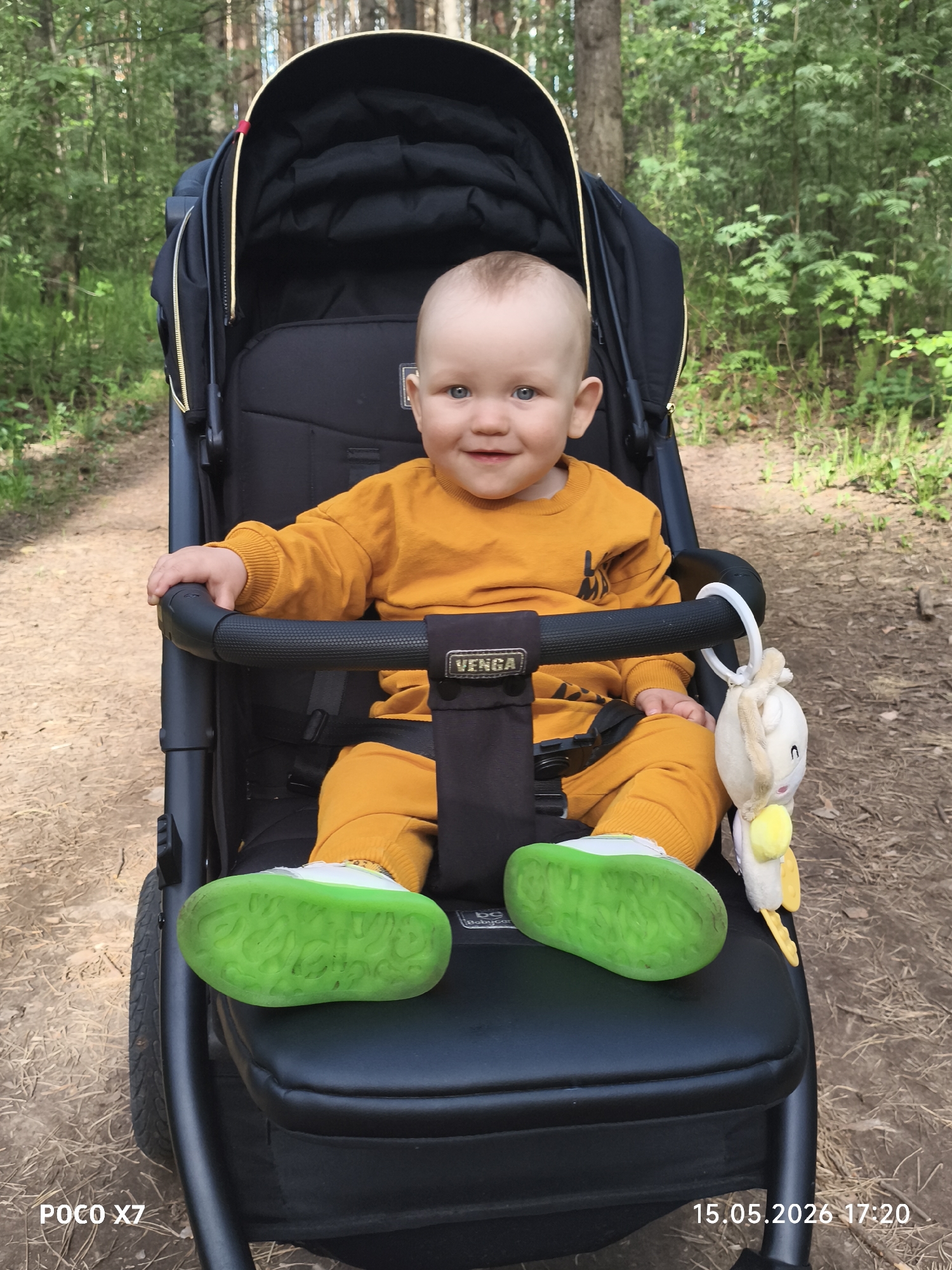 A baby sits in a black stroller in a forest. The baby wears a yellow outfit and white shoes with green soles. A plush toy hangs from the stroller's bar. The stroller is positioned on a dirt path amidst trees and foliage. The bottom left corner of the photo displays the text "POCO X7", while the bottom right corner shows "15.05.2026 17:20". The photo was likely taken in a wooded area in or near the city of Chaikovsky, Russia.