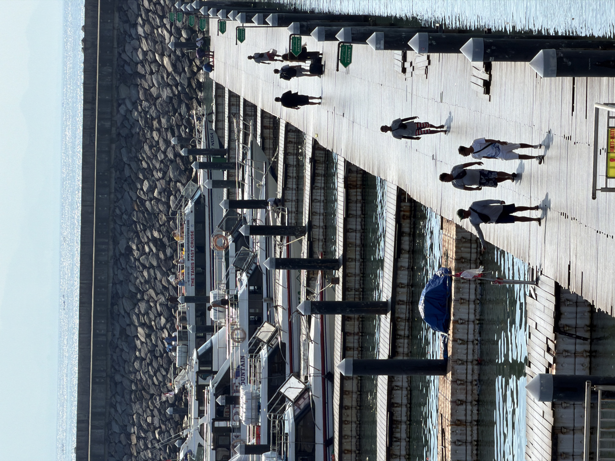 In Denpasar, Indonesia, a long wooden pier extends into a calm body of water, lined with numerous boats moored along its sides. The pier is made of weathered wooden planks and is supported by a series of dark, cylindrical posts. Several people are walking along the pier. In the foreground, a man wearing a t-shirt and striped shorts walks away from the camera. Further down the pier, a group of four men walk towards the camera. One man, in the lead, wears a t-shirt and shorts. Behind him, two men are walking side-by-side, one in a t-shirt and shorts, the other in a darker shirt and shorts. The fourth man, slightly behind, wears a t-shirt and shorts. Even further down the pier, two individuals, one carrying a dark suitcase, walk towards the camera. The background features a stone retaining wall separating the water from a hill, and beyond that, hazy mountains under a bright sky. The water is calm, reflecting the light, and several boats, including some with visible names like "NUSA" and "THE TANIS FAST CRUISE," are docked.