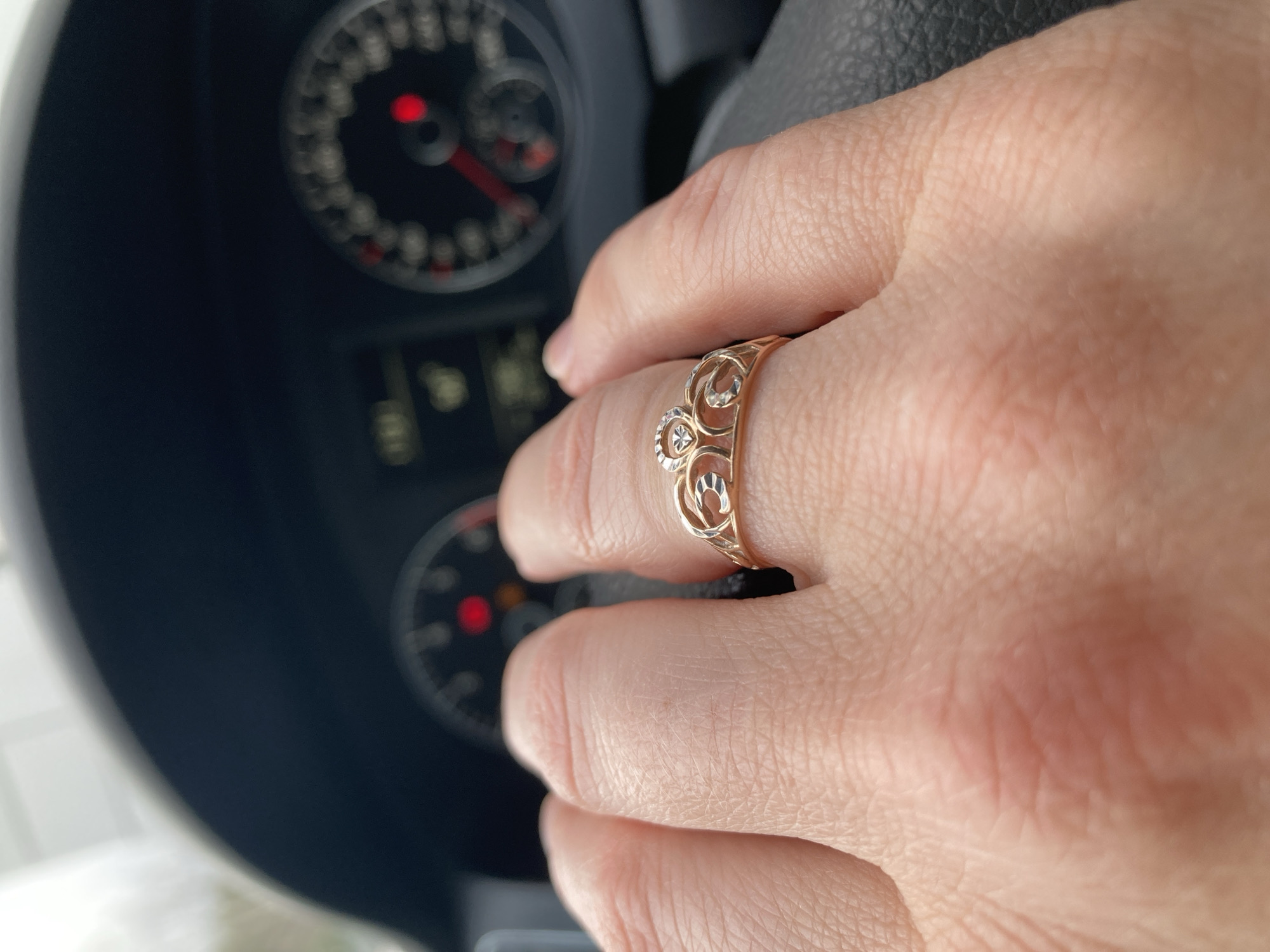 The image captures a close-up of a human hand, likely female, wearing a decorative ring, gripping a car's steering wheel. The hand, positioned in the lower-right portion of the frame, is well-lit, revealing skin texture and subtle red undertones. The ring, worn on the ring finger, is gold-toned with an intricate openwork design, featuring swirling patterns and three small, shiny elements that appear to be gemstones or polished metal accents.
In the background, out of focus, is the car's dashboard and instrument cluster. A speedometer or tachometer is partially visible on the left, displaying a red needle and a prominent red warning light. Other blurred indicators and parts of the dashboard are also discernible. The steering wheel, mostly covered by the hand, appears to be made of dark material, possibly leather, with visible stitching or texture.
The activity depicted is a person holding a steering wheel, suggesting they are either driving, parked, or a passenger in a car. The lighting indicates daylight conditions, but the exact time of day or weather cannot be determined. No emotions are visible, as only the hand is shown. There is no discernible text in the image due to the blur and angle. The scene is set inside a vehicle in Chaikovsky, Russia.