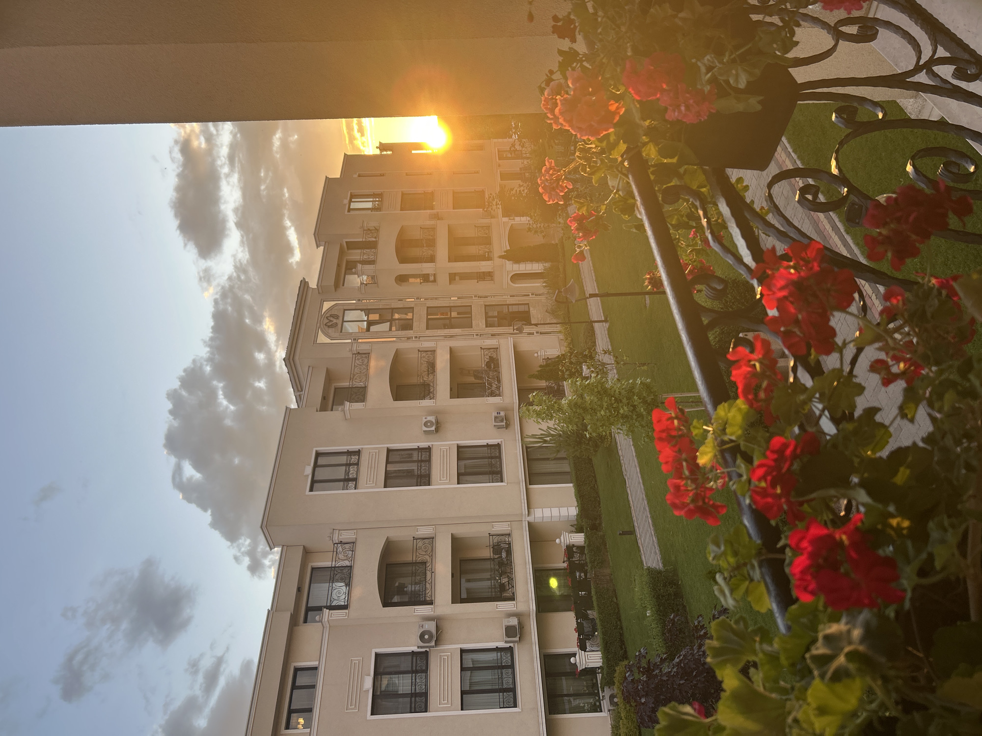 The image depicts a view from a balcony overlooking a modern apartment complex at sunset. The sun is setting on the right side of the frame, casting a warm, golden light across the scene. The sky is filled with dramatic clouds, tinged with orange and pink hues.
In the foreground, red geraniums in pots adorn the balcony railing, their vibrant color contrasting with the muted tones of the buildings. The apartment buildings are constructed with a light beige facade and feature multiple floors with balconies and windows. Air conditioning units are visible mounted on the exterior walls.
A well-maintained green lawn with manicured hedges and trees surrounds the buildings. A paved walkway winds through the grass, leading to the entrance of the complex. The overall scene suggests a peaceful and aesthetically pleasing residential environment. The architecture and landscaping are consistent with modern residential developments found in areas such as the Kapana district of Plovdiv, Bulgaria, which is known for its blend of historic charm and contemporary revitalization, often featuring similar apartment buildings with balconies and green spaces.