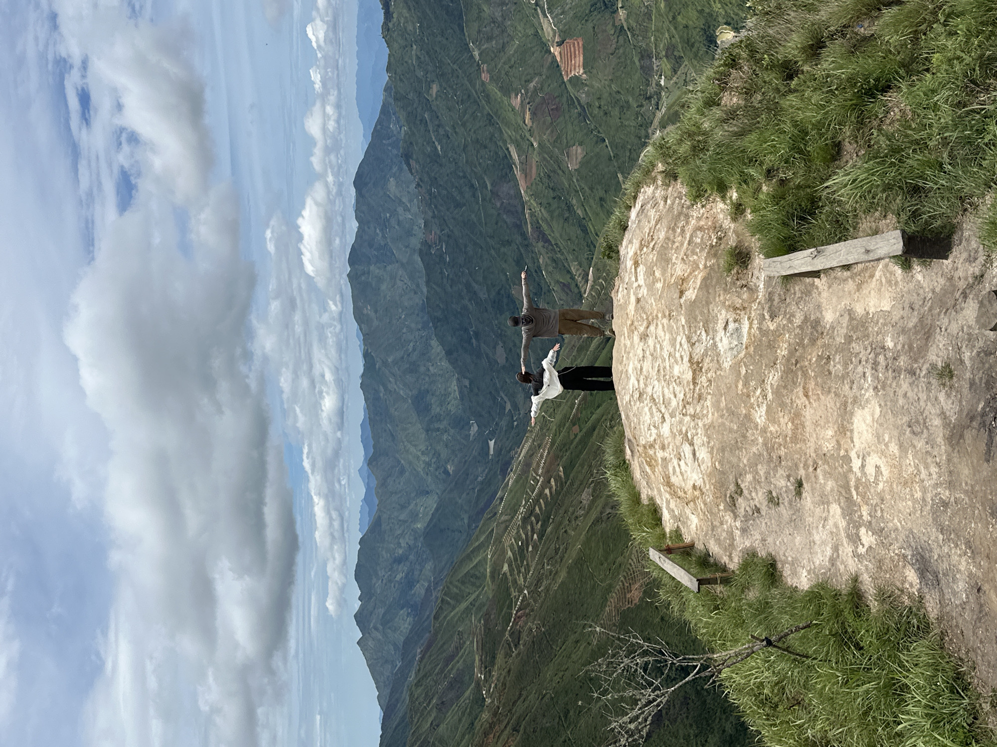 Two individuals stand on a rocky precipice, their arms outstretched in a gesture of embrace or freedom, overlooking a vast expanse of verdant, terraced mountains. The foreground is dominated by the rough, textured surface of the cliff, with patches of dry grass and a solitary, bare tree adding a touch of wildness. A simple wooden bench is visible on the left, indicating a designated viewing spot. The middle ground reveals the dramatic topography of the mountains, with winding roads and terraced fields carved into their slopes, suggesting human cultivation in harmony with the natural landscape. A river or valley snakes through the distant landscape, adding depth and a sense of scale. The background features a layered panorama of more mountains fading into the hazy distance, under a sky filled with dramatic, billowing clouds. This scene is characteristic of the breathtaking vistas found in Ta Xua, Son La province, Vietnam, known for its "cloud hunting" spots and rugged mountain terrain.