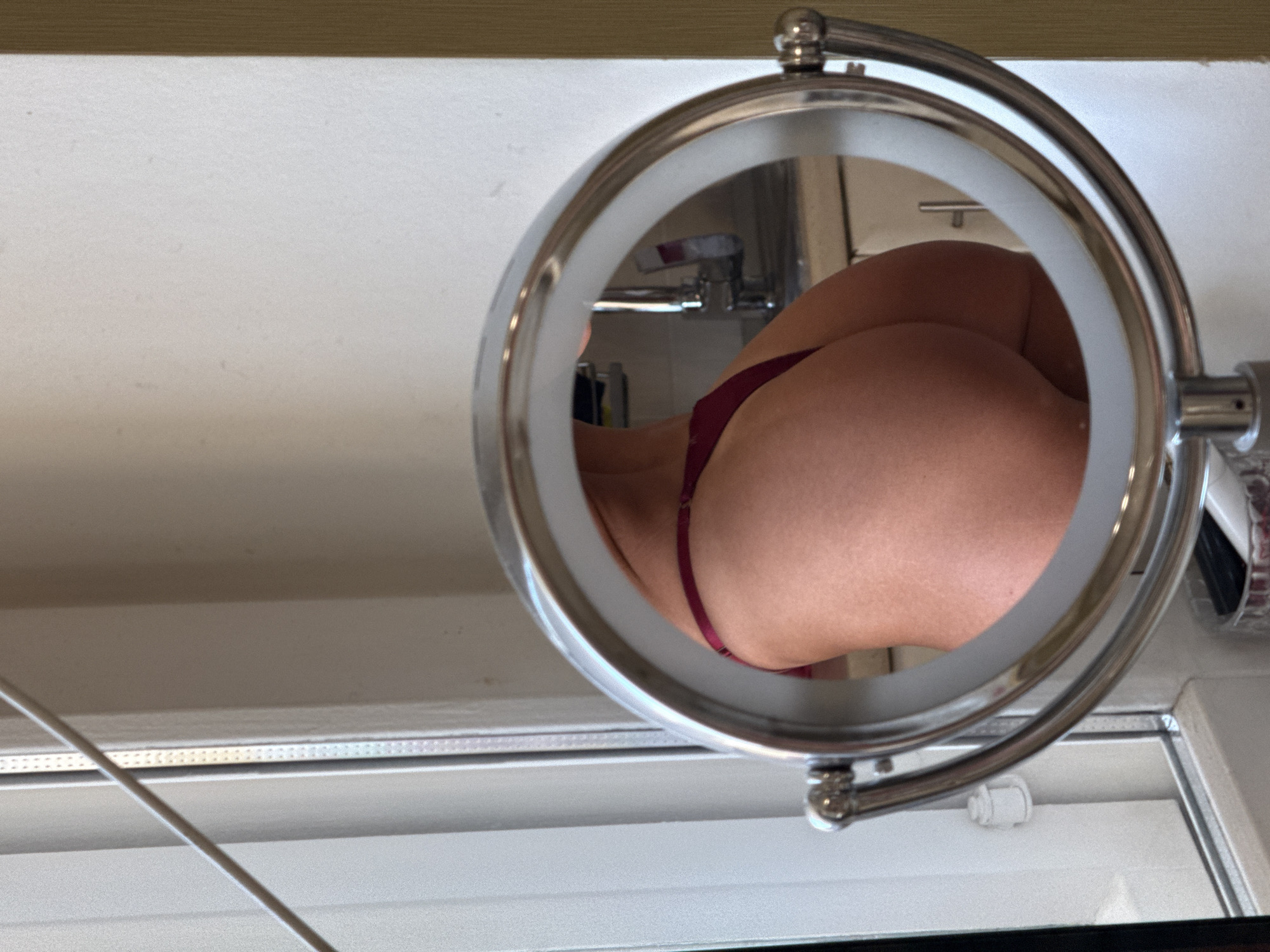 The media file shows a close-up shot of a person's buttocks reflected in a lighted makeup mirror. The reflection reveals the person is wearing burgundy underwear. The mirror is framed by a metallic, circular structure with a pivot mechanism. In the background, a faucet and sink are visible, suggesting a bathroom setting. The overall lighting appears to be bright, possibly from the mirror's integrated LED ring. No text is visible in the image. The scene is static, capturing a moment of self-reflection. The location context of Saint Petersburg, Russia, does not provide further visual cues within the image itself.
