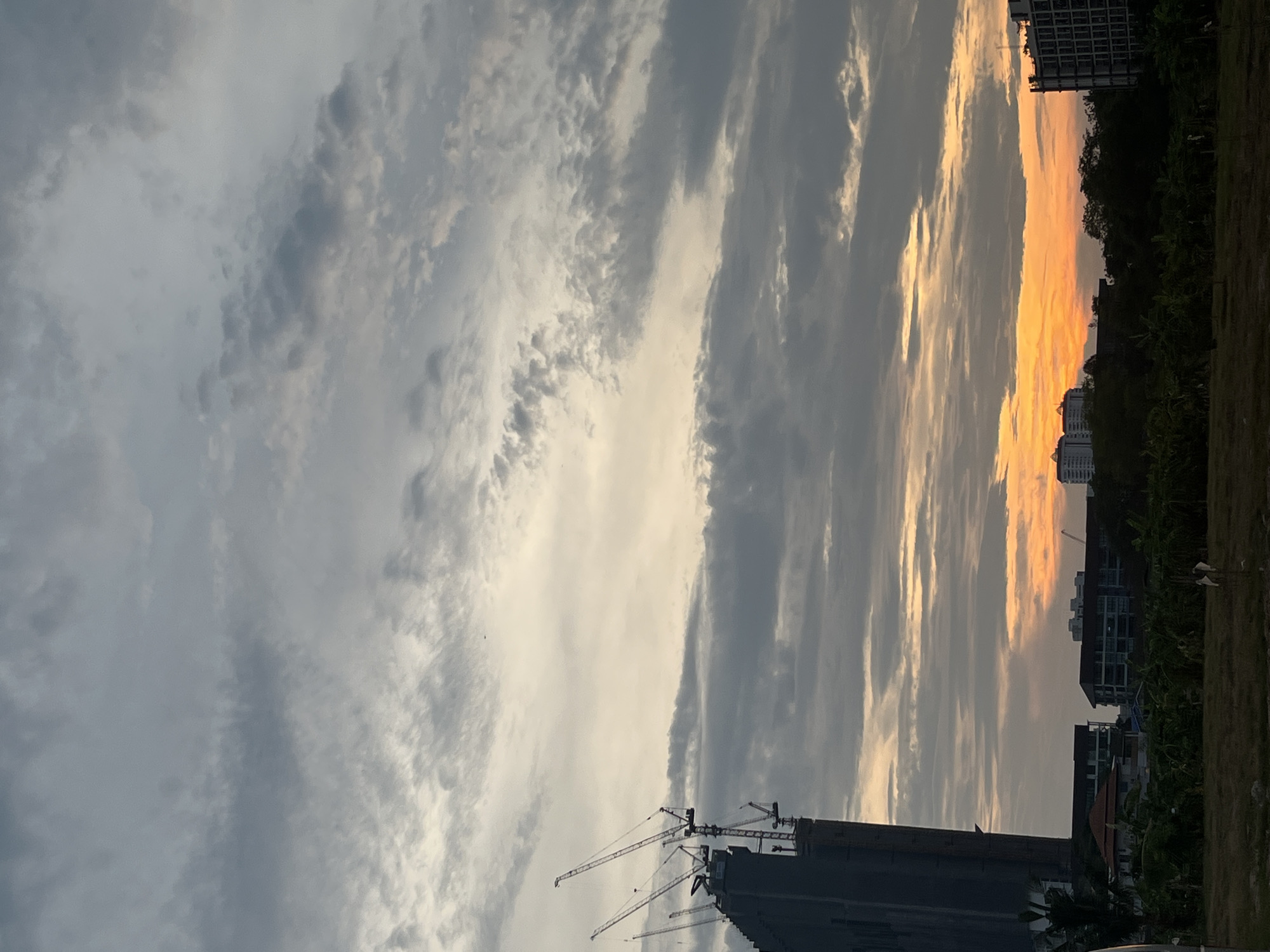 The image depicts a skyline in Pattaya City, Thailand during a sunset or sunrise. A grey building under construction stands in the foreground, with several cranes extending into the sky. The sky is filled with varying shades of grey and white clouds, illuminated by an orange or yellow glow from the rising or setting sun. The horizon line is lined with a mix of trees and buildings. In the distance, taller buildings can be seen. The scene suggests urban development within a natural landscape.