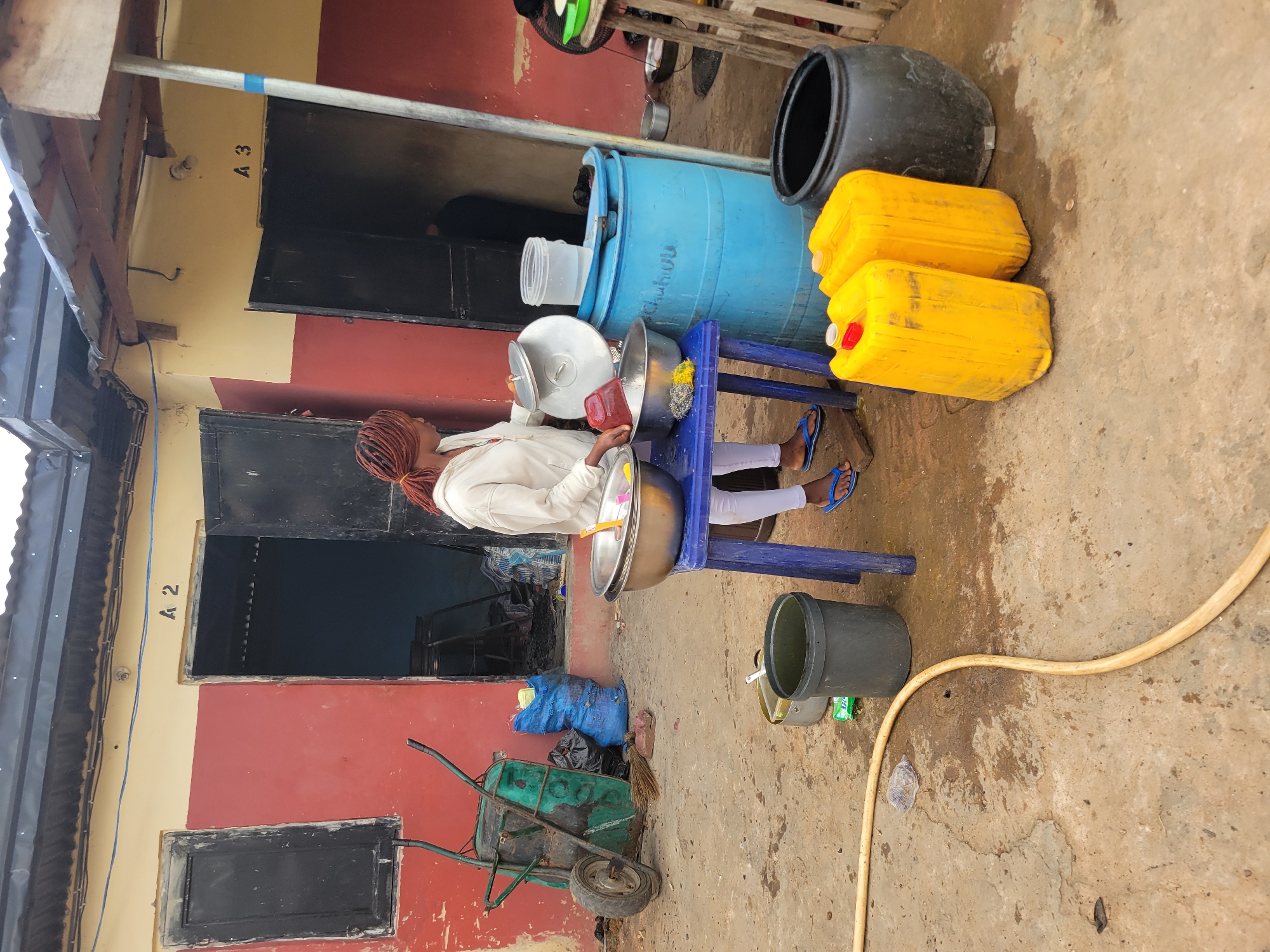 This is a medium-angle shot of an outdoor scene in Egbagu, Nigeria. The primary subject is a person, likely female, wearing a white hoodie and light-colored pants, standing and interacting with bowls on a blue table. The person has braided hair with reddish-brown highlights, tied back. She is wearing blue flip-flops.
The scene is cluttered with various objects. To the right of the person are two large, bright yellow jerrycans and a large black plastic barrel. Behind the person and the table is a large blue barrel. There are also metal bowls and pots on the table, some appearing to be for cooking or cleaning. A broom and a green wheelbarrow are visible on the ground to the left of the person. A yellow hose lies on the ground, snaking across the frame. The ground is a dirt or concrete surface, appearing somewhat damp or dirty. In the background, there is a red and yellow building with open doorways.
The activity appears to be preparation or cleaning, given the bowls and the presence of a wheelbarrow and broom. The time of day is not explicitly indicated, but the lighting suggests it is daytime. The weather seems overcast, as there are no strong shadows. The location cues, such as the building style and the objects present, are consistent with a rural or semi-urban setting in Nigeria. There is some visible text: "A 3" is written on the yellow wall to the left, and "A 2" is written on the wall above a dark rectangular opening on the left side of the image.