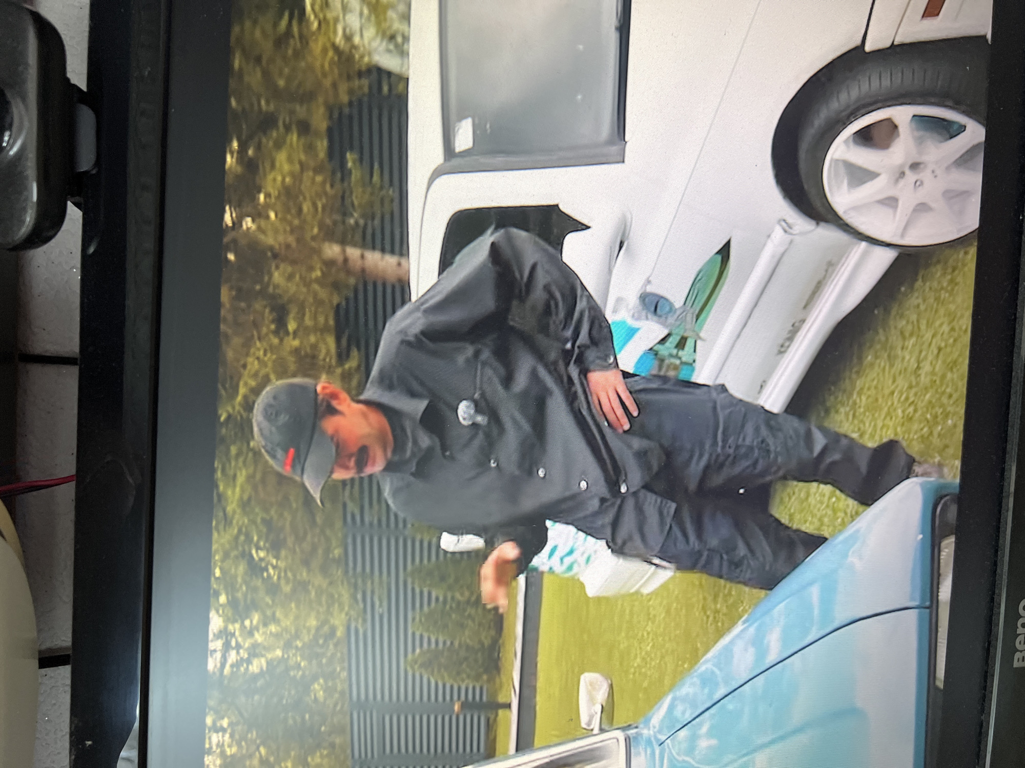 A man wearing a black button-down shirt, black cargo pants, and a black baseball cap with a red logo stands between two cars on a grassy lawn in front of a grey metal fence. The man has a mustache and is gesturing with his right hand. To his left is the front fender and headlight of a light blue car. To his right is the side of a white car with a white racing stripe and the word "KONIG" visible on the rocker panel. The white car has a white multi-spoke rim with a black tire. The background shows green trees and bushes. The image appears to be a photograph taken of a screen. The location is identified as Kanash, Russia.