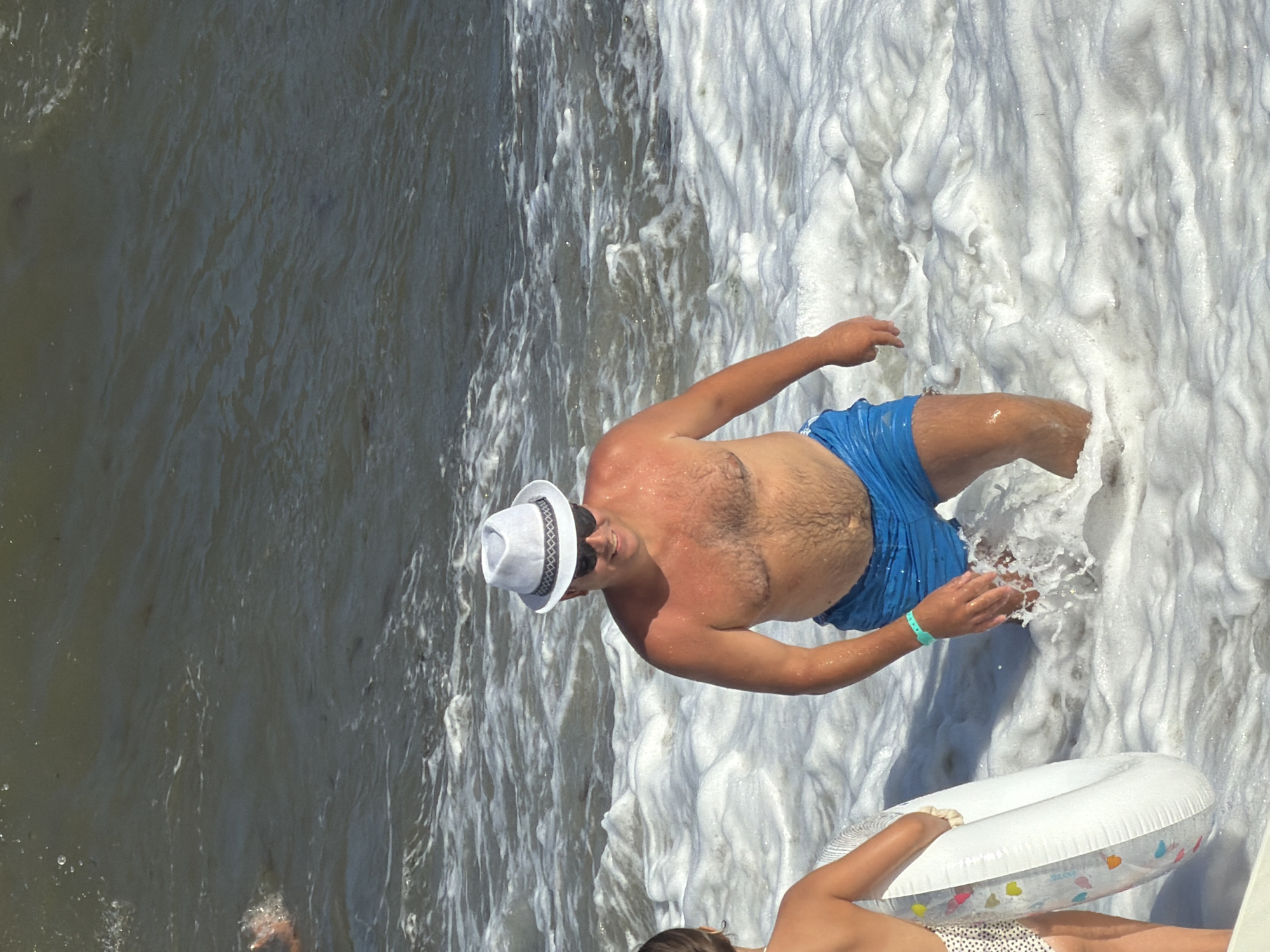 The image depicts a man in blue swim trunks and a white fedora hat standing in the ocean waves. He is wearing sunglasses and a green bracelet on his left wrist. To his left, a child is partially visible, holding a white inflatable ring with colorful heart designs. The water is choppy with white foam, and the overall scene suggests a beach setting. The image is not geotagged and there is no information to suggest the location is in Alushta, Ukraine.