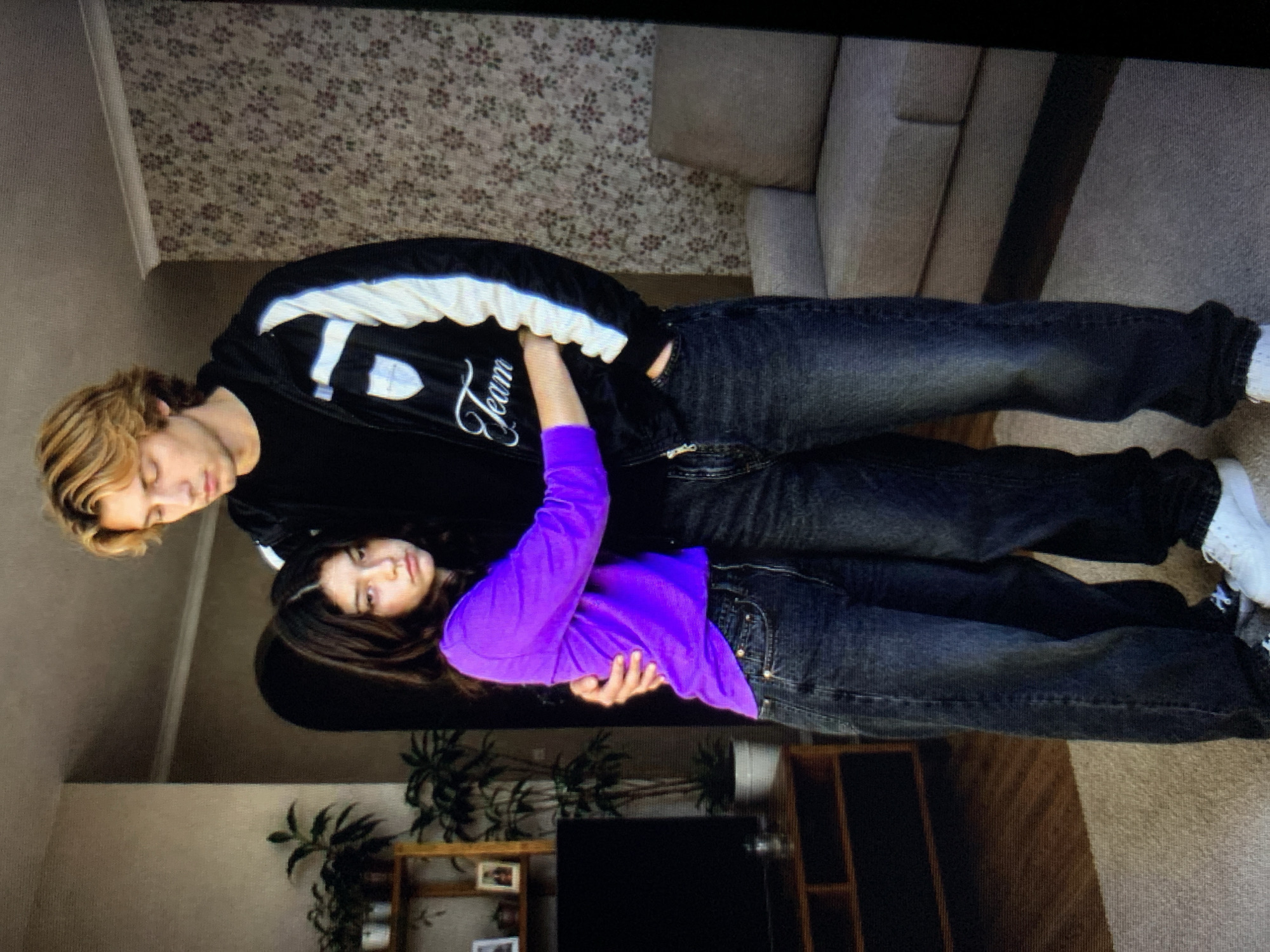 A young man and woman stand in a room with floral wallpaper and a grey sofa. The man, who is taller than the woman, is wearing a black t-shirt, a black and white jacket with the word "Team" on it, and dark jeans. He has blonde hair and is looking down. The woman is wearing a bright purple long-sleeved shirt and dark jeans. She is embracing the man and looking at the camera with a sad expression. In the background, there is a bookshelf with plants and picture frames, and a television. The scene appears to be indoors, possibly in a residential setting in Lymanka, Ukraine.