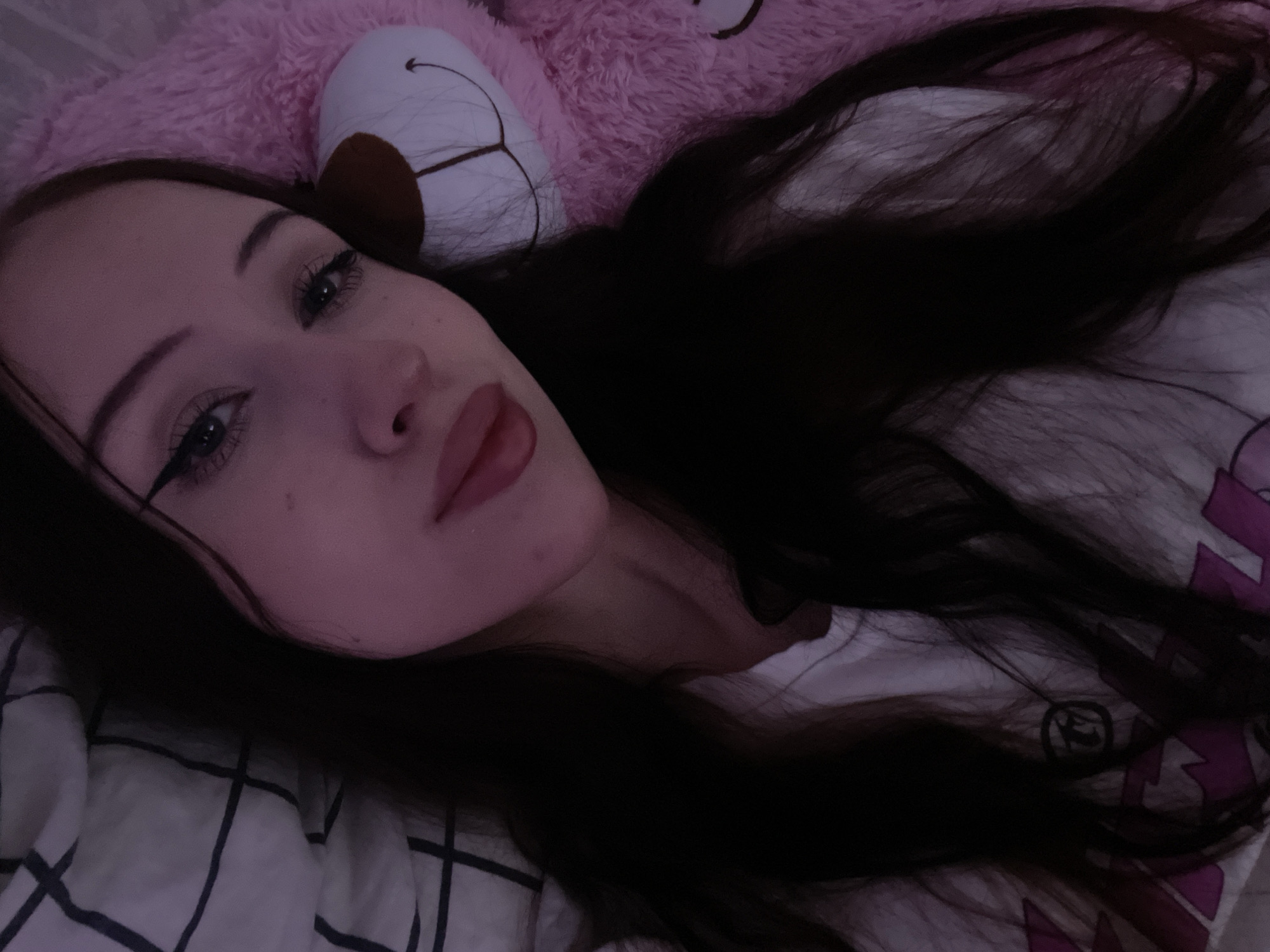 The image shows a young woman with long, dark hair, lying down on a bed. She is wearing a white t-shirt with a graphic design. To her right, a large, pink stuffed bear with a smiley face is visible. The background includes a striped pillow and a pink fuzzy blanket. The lighting in the image is dim, creating a soft, diffused effect. There is no discernible connection to Votkinsk, Russia, or any specific urban environment. The scene appears to be an indoor, personal space, likely a bedroom. The woman is looking towards the camera, with a neutral expression. The image is a close-up, focusing on her face and upper body.