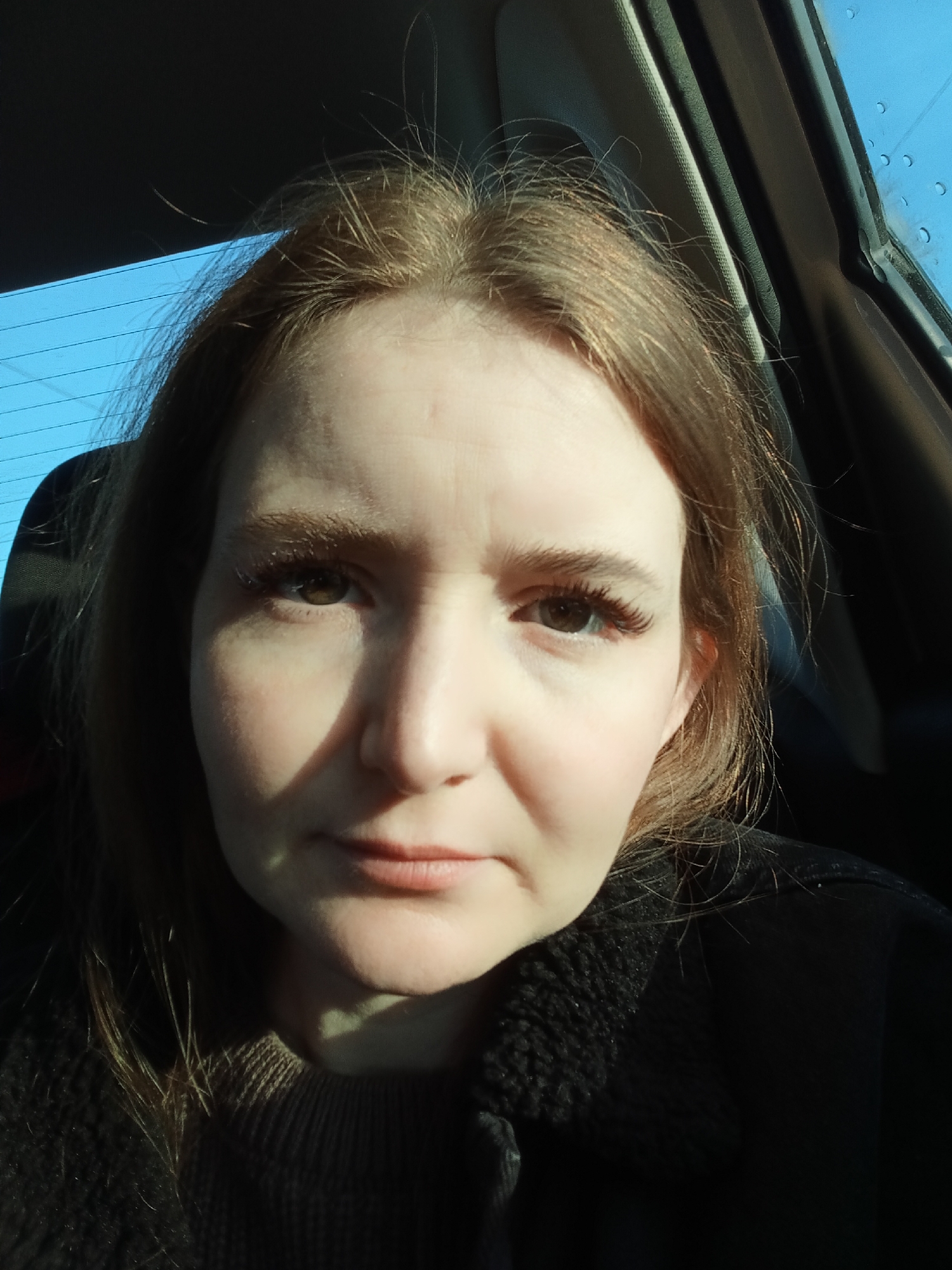 A medium close-up selfie photograph shows a fair-skinned woman with light brown hair, brown eyes, and false eyelashes. She is looking directly at the camera with a neutral expression. She is wearing a brown sweater and a dark black jacket with a textured collar. The setting appears to be inside a car, as suggested by the window with condensation droplets, the car seat, and the window frame visible in the background. A bright blue sky can be seen through the window. The location, Chaikovsky, Russia, cannot be definitively confirmed based on the image alone.