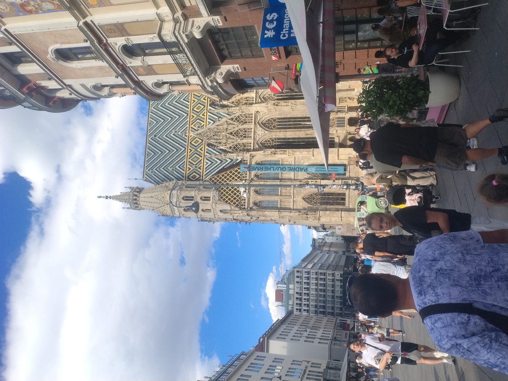 The image depicts a crowded pedestrian area in Vienna, Austria, likely Stephansplatz, given the presence of St. Stephen's Cathedral, recognizable by its distinctive patterned roof and Gothic architecture. The cathedral's spire dominates the background, while the surrounding buildings showcase a mix of historical and modern styles.
In the foreground, several individuals are walking or standing. A man in a blue patterned shirt is seen from behind, with a black strap across his shoulder. To his right, a young woman in a black t-shirt and white shorts looks towards the camera. Further back, a man in a white polo shirt and black shorts walks away from the camera, holding a phone. Another man in a black t-shirt and shorts is walking towards the right side of the frame.
Numerous other people are dispersed throughout the scene, engaged in various activities such as walking, browsing, or sitting at outdoor cafes. A green vintage car is visible in the middle ground, adding a touch of color to the scene. Several umbrellas and awnings suggest outdoor dining or market stalls. The sky is a mix of blue with scattered white clouds, indicating fair weather. Signs for "Change" and currency symbols (Yen, Euro, Dollar) are visible on a building to the right, suggesting a currency exchange service.