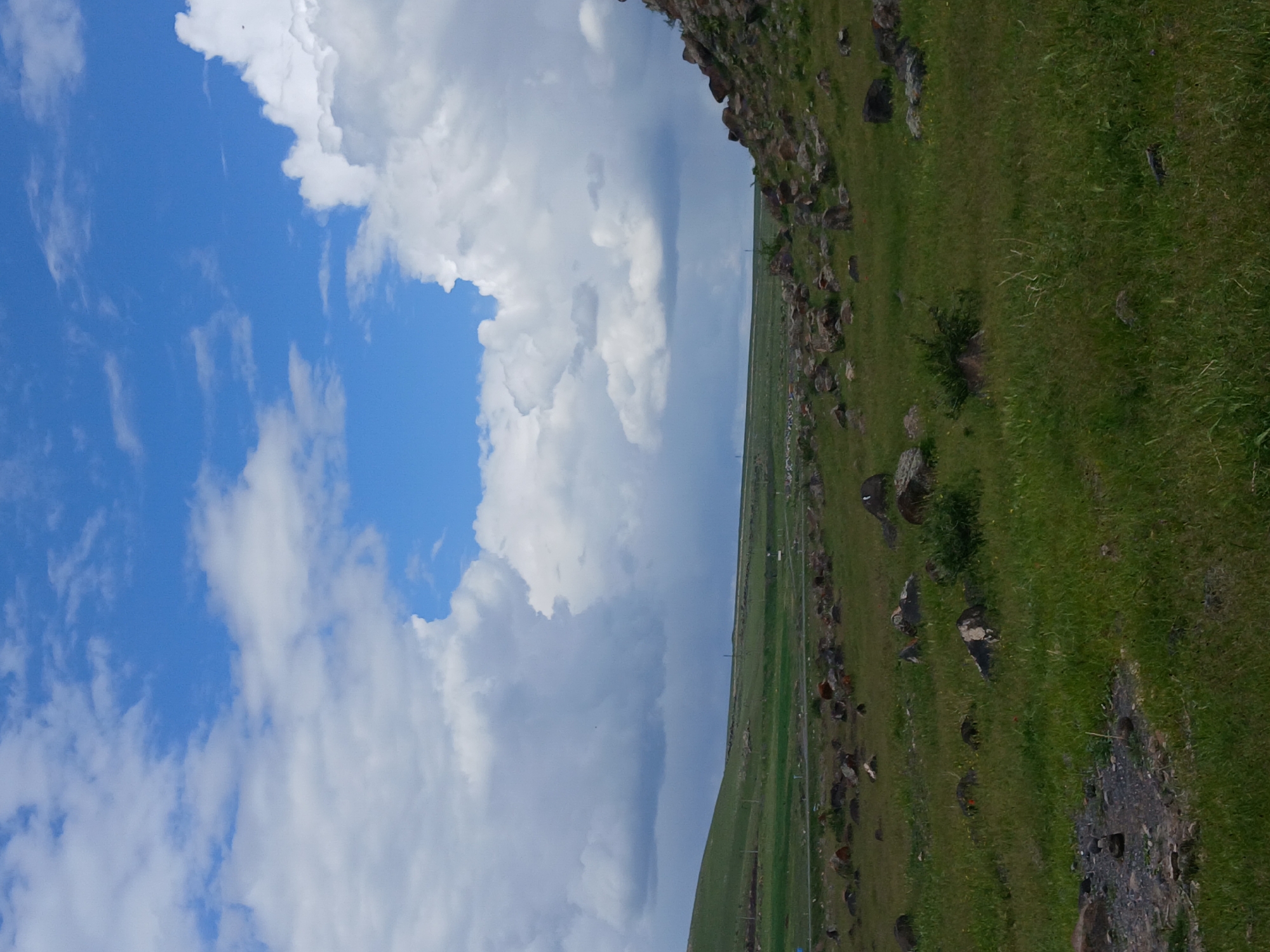 The image shows a landscape near Tlik, Armenia. The left side features a blue sky with white cumulus clouds. The right side shows a green hillside with scattered rocks. In the middle ground, there is a flat expanse of green land. The image appears to have been taken from a high vantage point, possibly overlooking the terrain. There are no people visible in the frame. The main subjects are the sky, clouds, hillside, and grassy plain. The environment is rural and mountainous. The action is stillness, a static scene.