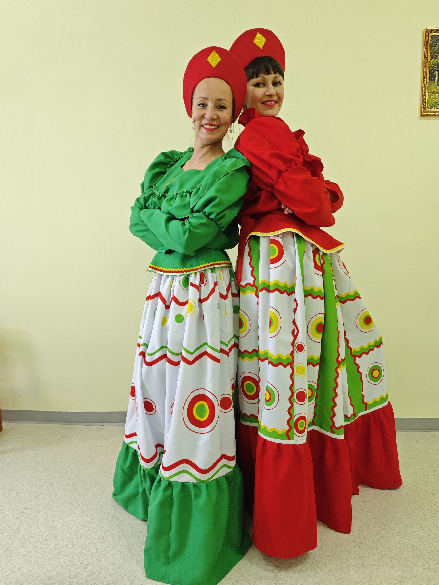 Two women, smiling brightly, stand back-to-back with their arms crossed in a full-length portrait. They are dressed in vibrant, traditional-style costumes. The woman on the left wears a bright green, long-sleeved blouse with ruffled details and a long, full white skirt adorned with concentric red, green, and yellow circles, and wavy lines. Her skirt has a green ruffled hem. She wears a red hat, similar to a kokoshnik, featuring a yellow diamond-shaped embellishment.
The woman on the right wears a similarly styled bright red, long-sleeved blouse with ample ruffles, and a matching long white skirt with the same circular and wavy line patterns in red, green, and yellow. Her skirt finishes with a red ruffled hem. She also wears a red hat with a yellow diamond-shaped accent.
They are positioned against a plain, light yellow wall. The floor is a light, neutral color, possibly linoleum or carpet. In the upper right corner of the background, a small, framed landscape painting is visible on the wall. The setting appears to be indoors, likely a community hall, school, or cultural center in Markovskii, Russia. Both women exude a joyful and friendly demeanor, suggesting they are either performing or posing for a cultural event or photoshoot. There is no visible text.
