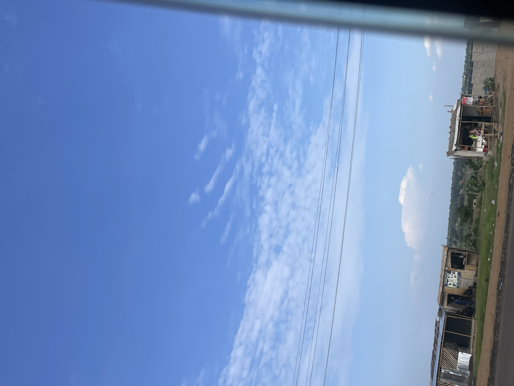 This image, captured in Uke, Nigeria, depicts a daytime roadside scene under a clear blue sky with scattered cumulus clouds. Multiple dark power lines stretch horizontally across the middle ground.
On the left, two adjacent, open-fronted, single-story structures with corrugated metal roofs and wooden frames are visible. The structure on the far left appears unfinished or in disrepair, with visible wooden scaffolding and a partial corrugated metal roof. The structure immediately to its right is more complete, featuring walls that appear to be a mix of wood planks and possibly sheet metal. Several individuals are present in and around these structures; at least two people are seated outside the second structure, and others are partially obscured inside. Signs with text and images are affixed to the front of this structure.
Further to the right, a third, smaller, single-story roadside stall is visible. This stall also has a corrugated metal roof and appears to be constructed from wooden planks. It features an open counter area where goods are displayed, and at least one person is visible behind the counter. A red sign with white text, partially reading "POS," is attached to the front right side of this stall.
The foreground consists of a dirt and gravel road shoulder with sparse green vegetation and scattered debris. Beyond the structures, the landscape recedes into rolling hills covered with greenery under the distant sky. A concrete or stone wall is partially visible behind the rightmost stall. A dark vertical element on the far right edge of the frame suggests the image was taken from inside a vehicle, possibly through a window frame.