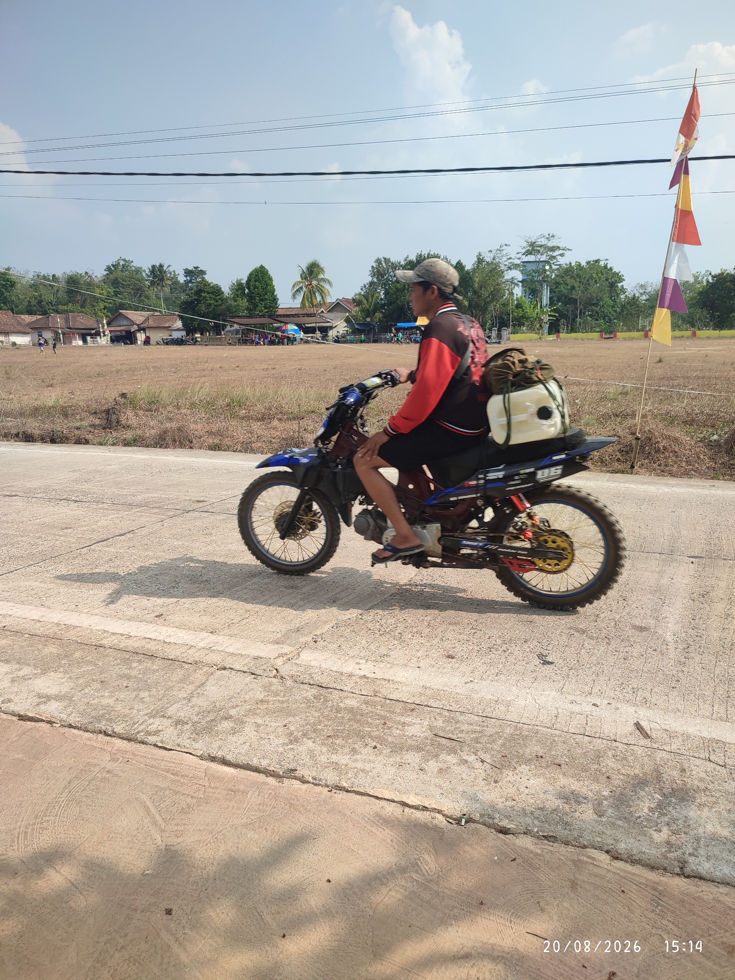 A man wearing a cap, red and black jacket, and shorts rides a blue and black dirt bike on a paved road. He has a light-colored backpack attached to the rear of the motorcycle. The road is bordered by dry, brown grass and a dusty field. In the background, there are several small buildings with brown roofs, trees, and a tall water tower. A pole with several colorful flags, red, white, yellow, and purple, stands to the right of the motorcycle. The sky is blue with some clouds. The timestamp "20/08/2026 15:14" is visible in the lower right corner. The scene appears to be located in Nusa Jaya, Indonesia, based on the provided information.