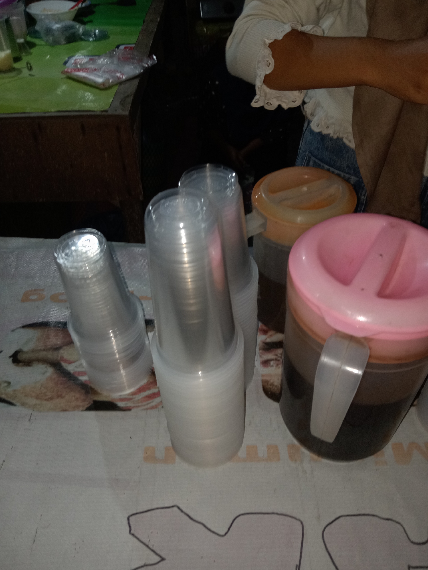 The image shows a close-up view of disposable plastic cups stacked neatly, positioned in the foreground. Behind the cups, two large beverage dispensers are visible; one has a pink lid and the other an orange lid, both filled with a dark liquid. The dispensers are placed on a table covered with a patterned cloth. In the background, a person's arm wearing a white, ruffled top and denim jeans is partially visible, suggesting they are interacting with the items on the table. The environment appears to be a casual setting, possibly a food stall or a market in Stiaji, Indonesia, given the common use of such disposable cups and dispensers in these areas.