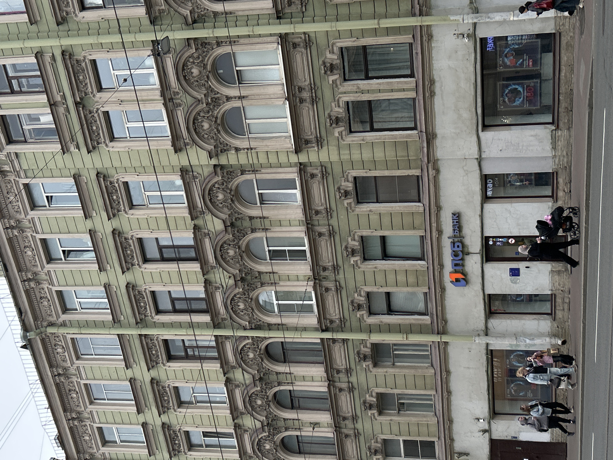 The image shows a multi-story building in what appears to be Saint Petersburg, Russia. The building's facade is adorned with ornate architectural details, featuring a pale green color scheme and multiple windows. Some windows are arched. A street is visible in the foreground, with pedestrians walking along the sidewalk. A sign with Cyrillic characters is visible on the building indicating "PSB Bank" which is Promsvyazbank. A group of people walks past what looks like a bank's facade. There are visible power lines running along the building. The weather seems overcast.
