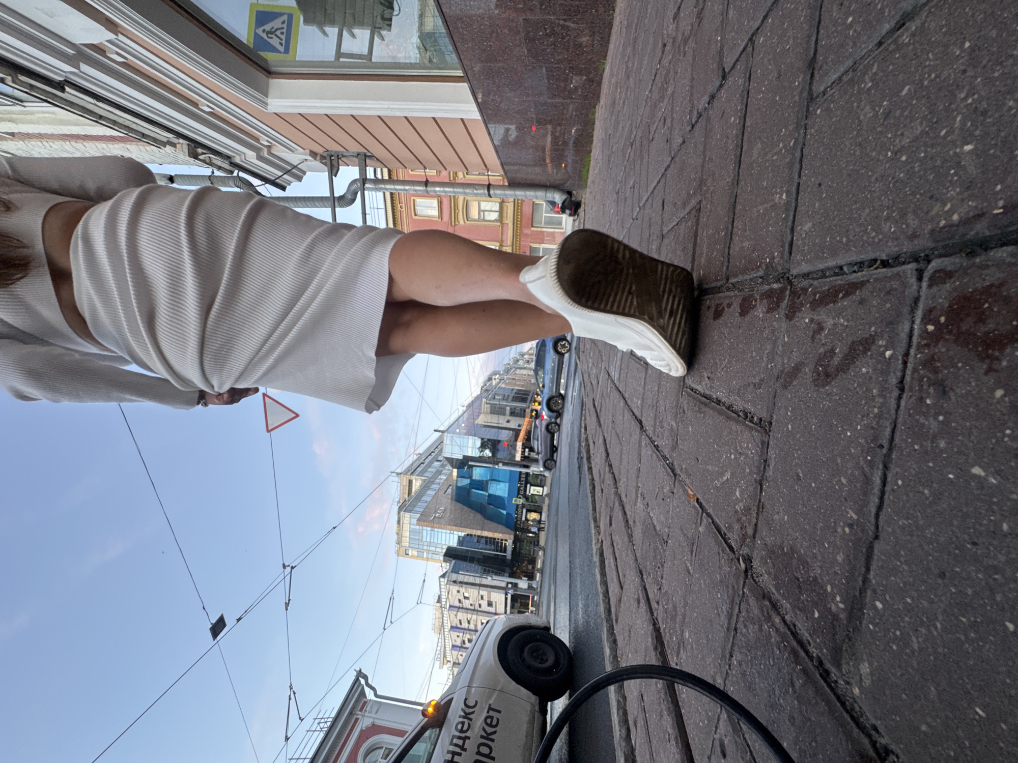 A low-angle, medium shot captures the lower body of a person walking away from the camera on a brick-paved sidewalk in Nizhny Novgorod, Russia. The person is wearing a white ribbed knit skirt and white sneakers. The skirt is high-waisted and hugs the curves of the wearer's legs. The sneakers appear worn and dirty. The sidewalk is made of dark gray rectangular bricks.
In the background, a white vehicle with Cyrillic lettering "Яндекс маркет" (Yandex Market) is parked at the side of the road. Beyond the car, modern glass-fronted buildings rise against a pale blue sky with wisps of pink clouds. Overhead, a network of tram or trolleybus wires crisscrosses the sky. A red and white triangular yield sign is visible on a pole to the right of the person. The overall lighting suggests either early morning or late afternoon.