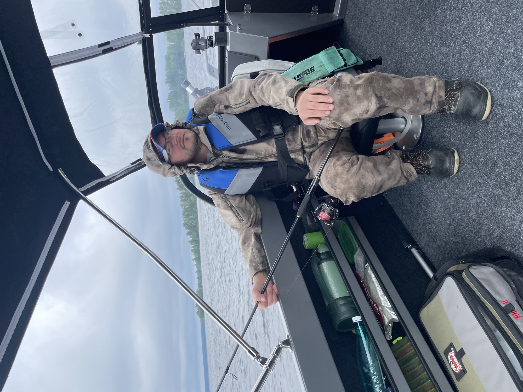 The image depicts a man sitting in a boat, steering. He is wearing camouflage clothing, a blue and grey life vest, glasses, and a hooded jacket. The boat appears to be on a body of water, with trees and a cloudy sky visible in the background. Inside the boat, there are fishing rods, tackle boxes, and a water bottle. The overall scene suggests a fishing excursion. The location is identified as Yablonovka, Russia.
