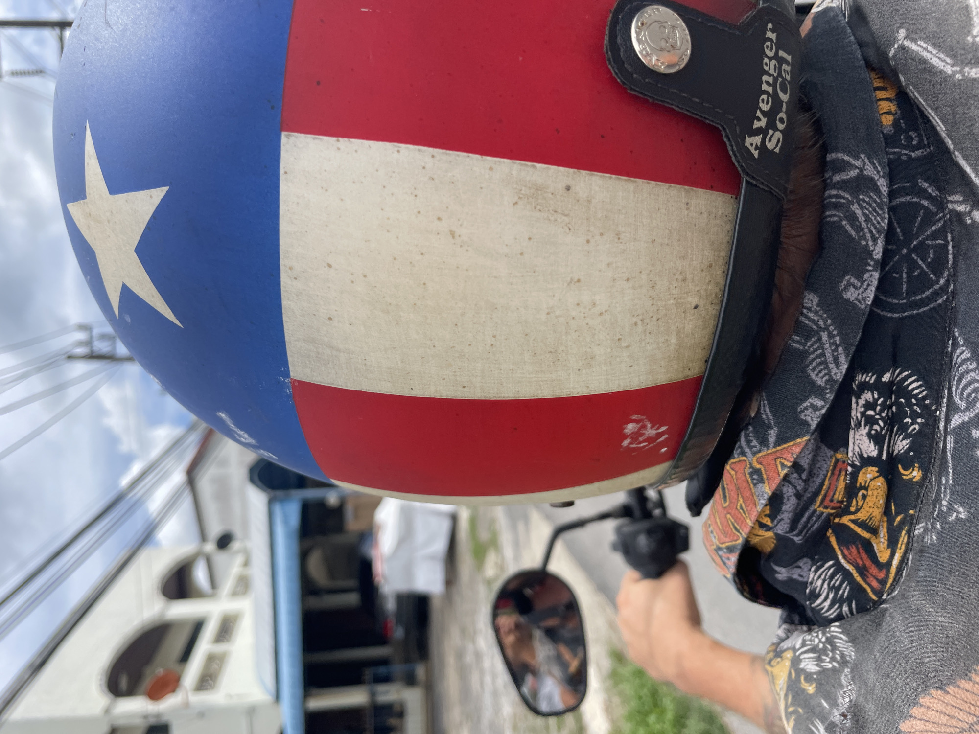 A close-up, eye-level shot depicts a motorcycle helmet painted with the colors of the Texas state flag—blue with a white star and red and white stripes. The helmet is branded "Avenger So-Cal." A person, partially visible, wears a patterned shirt or bandana. A side mirror reflects a blurry image of a person. The surrounding environment appears to be an urban street, likely in Choeng Thale, Thailand, though specific landmarks are obscured. Elements of architecture and power lines are also visible in the background. The photo seems to be taken from the perspective of the motorcycle rider.