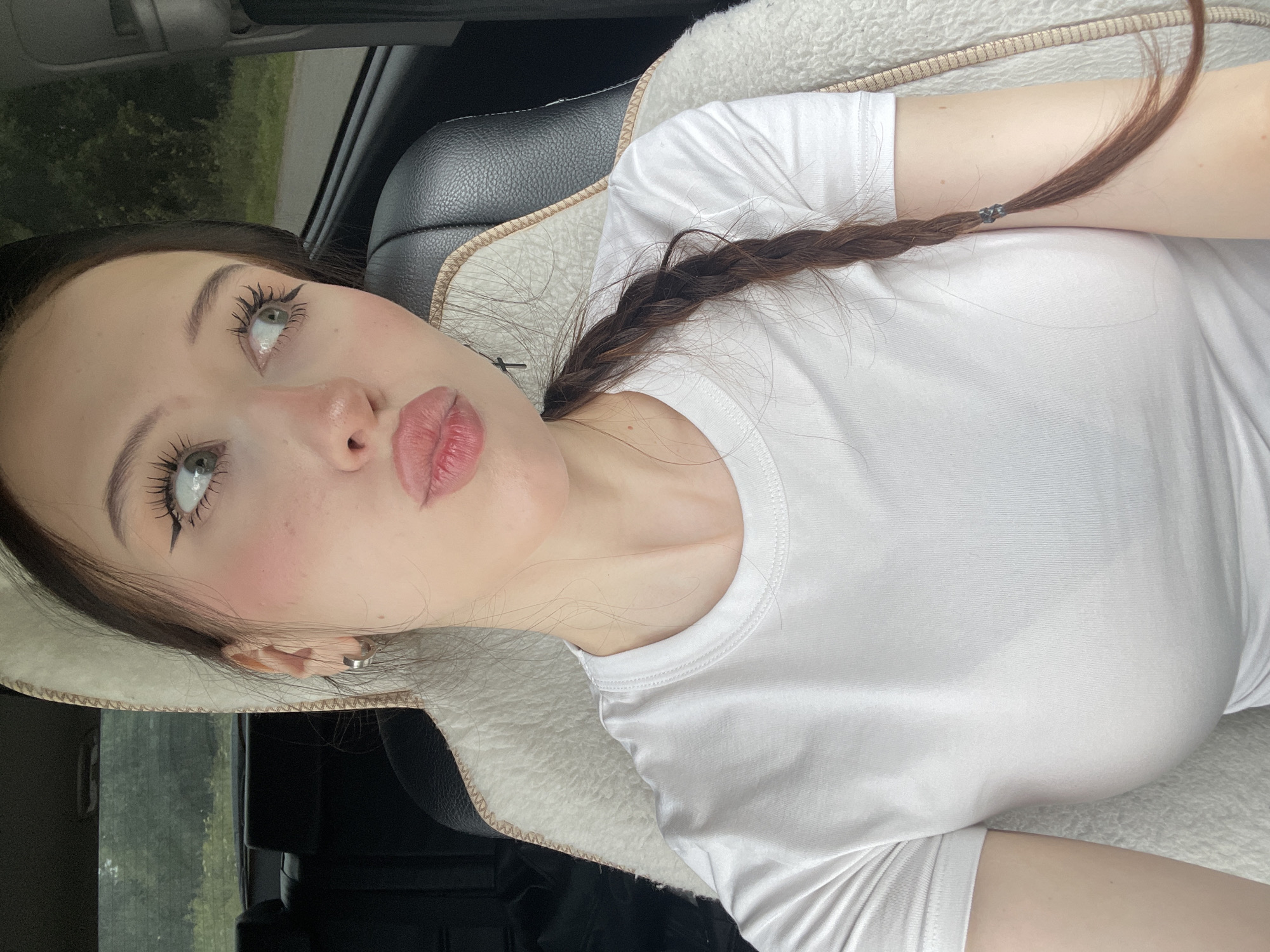 A young woman with light skin, dark brown hair braided to her right, and green eyes is sitting in the driver's seat of a car. She is wearing a white t-shirt. She is looking to her right and her lips are slightly pursed. The interior of the car is visible, including the headrest and a fluffy white seat cover. The background outside the car windows shows trees and foliage, suggesting a rural or suburban setting. There are no discernible landmarks to indicate the specific location within Gavrilovka, Russia.